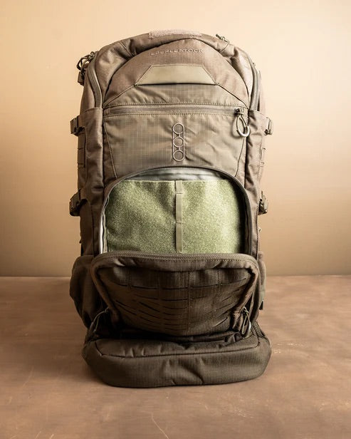 Der taktische Rucksack Spectre von EBERLESTOCK in Khakigrün steht aufrecht auf einer braunen Fläche, wobei das vordere Fach geöffnet ist und das grüne Innere zum Vorschein kommt. Er verfügt über mehrere Riemen, mehrere Taschen und ein schlankes, minimalistisches Design.