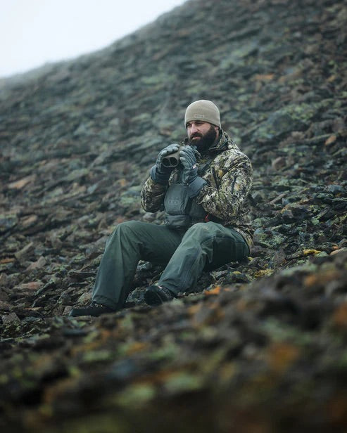 Ein bärtiger Mann in Tarnjacke, olivfarbener Hose und einer EBERLESTOCK North Fork Merino Beanie sitzt an einem bewölkten Tag auf einem felsigen Hügel, hält ein Fernglas in der Hand und begutachtet das Terrain.