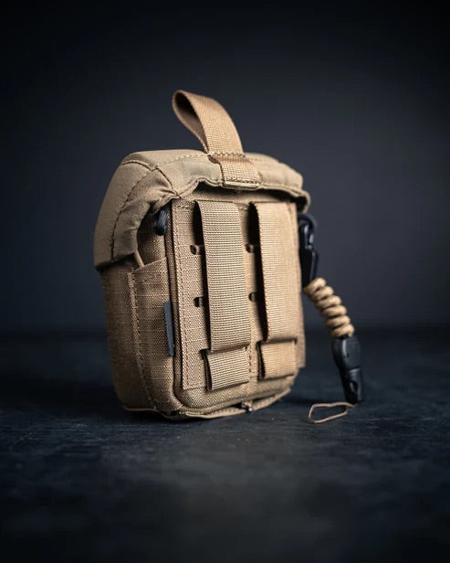 Die EBERLESTOCK Mag-Free Rangefinder Pouch, eine kleine, hellbraune Tasche mit MOLLE-Gurten und einer Zuglasche, steht aufrecht auf einer dunklen Oberfläche und ist mit einem aufgerollten Lanyard versehen - ideal für Eberlestock- oder Jagd Outdoor-Taschen-Setups.