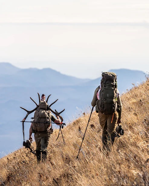 Zwei Wanderer mit großen Rucksäcken und Trekkingstöcken gehen einen trockenen Hang hinauf; einer trägt ein Hirschgeweih. Zu ihrer Ausrüstung gehört der Rucksack EBERLESTOCK Brute 6500. Im dunstigen Hintergrund erheben sich Berge.