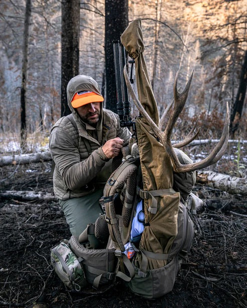 Ein Mann in Outdoor-Ausrüstung kniet in einem verbrannten Wald und rückt seinen EBERLESTOCK Brute 4500 Rucksack zurecht, der mit Elchgeweihen und einem Gewehr beladen ist. Er trägt eine orangefarbene Mütze und eine hellbraune Jacke inmitten von heruntergefallenen Ästen.