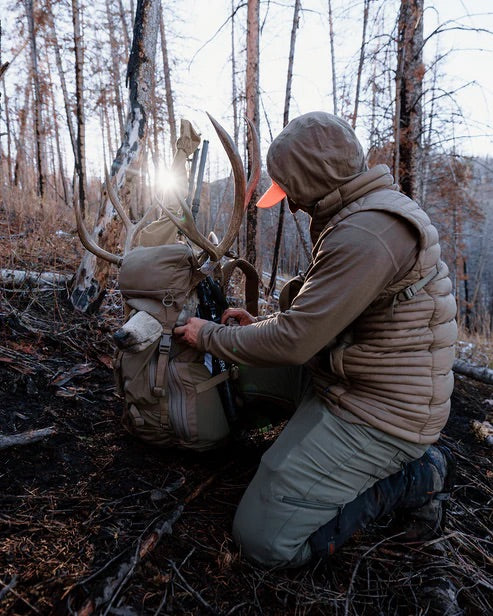 Eine Person in Outdoor-Ausrüstung kniet in einem Wald und justiert einen EBERLESTOCK Brute 4500 Rucksack mit einem Hirschgeweih darauf. Das Sonnenlicht fällt durch die Bäume und erzeugt einen hellen Fleck hinter dem Geweih.