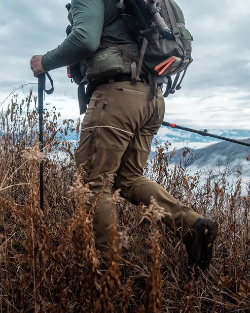 Eine Person mit der Eberlestock Deadwood Pant - Coyote Brown von EBERLESTOCK und einem Rucksack wandert einen grasbewachsenen Hügel hinauf, mit Trekkingstöcken, Bergen und einem bewölkten Himmel im Hintergrund.