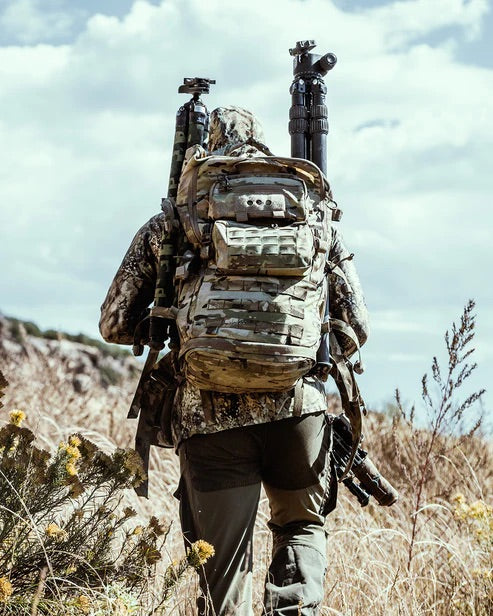 In Tarnkleidung geht eine Person mit einem EBERLESTOCK Mission Ruck und Stativen durch hohes Gras in zerklüftetem Gelände unter einem bewölkten Himmel.