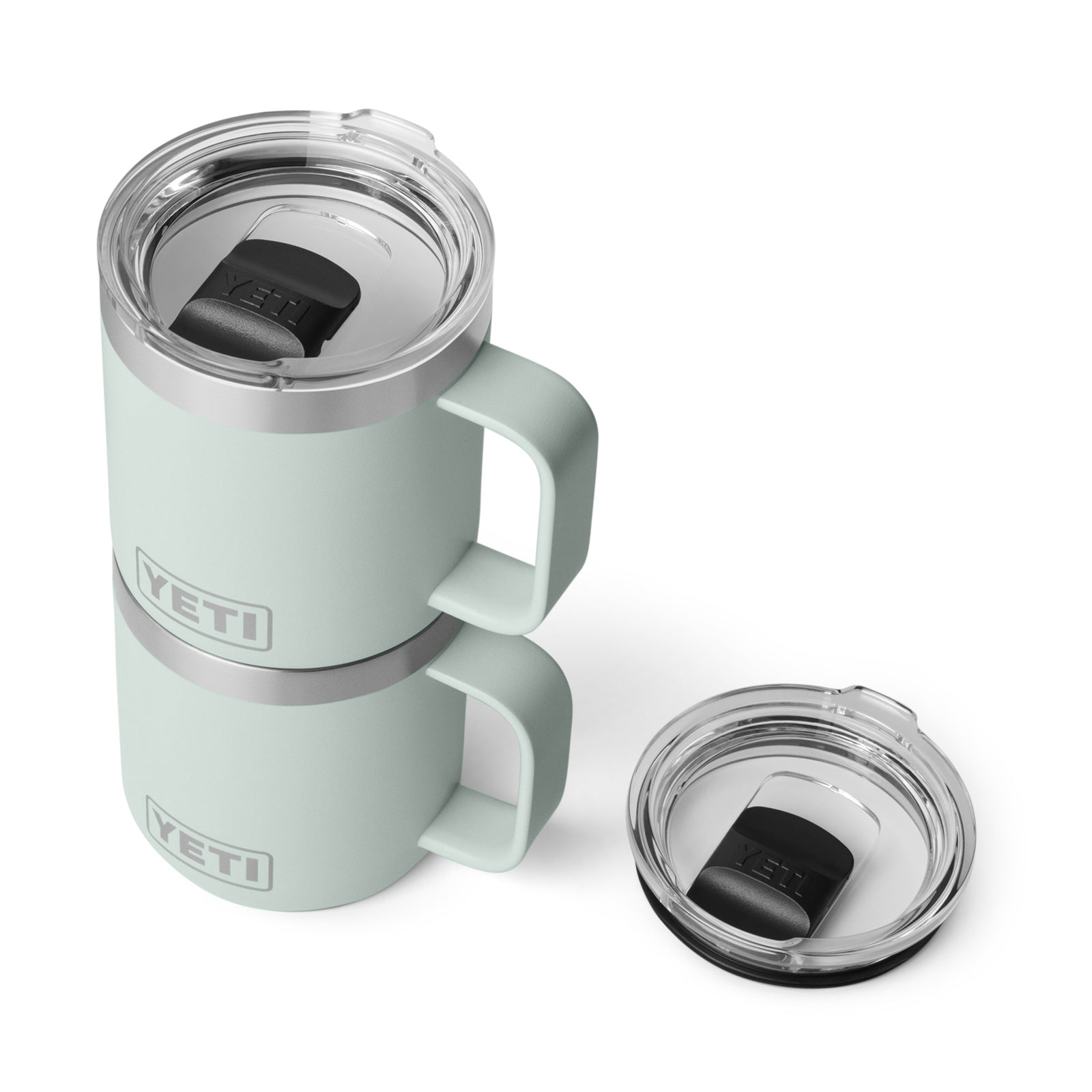 Zwei hellgrüne YETI® Tasse Rambler 10 Oz - Ridgeline Becher mit Griffen und durchsichtigen Deckeln sind gestapelt, mit einem zusätzlichen Deckel daneben. Die Becher haben auffällige YETI-Logos und eine doppelwandige Vakuum-Isolierung für eine hohe Temperaturstabilität.