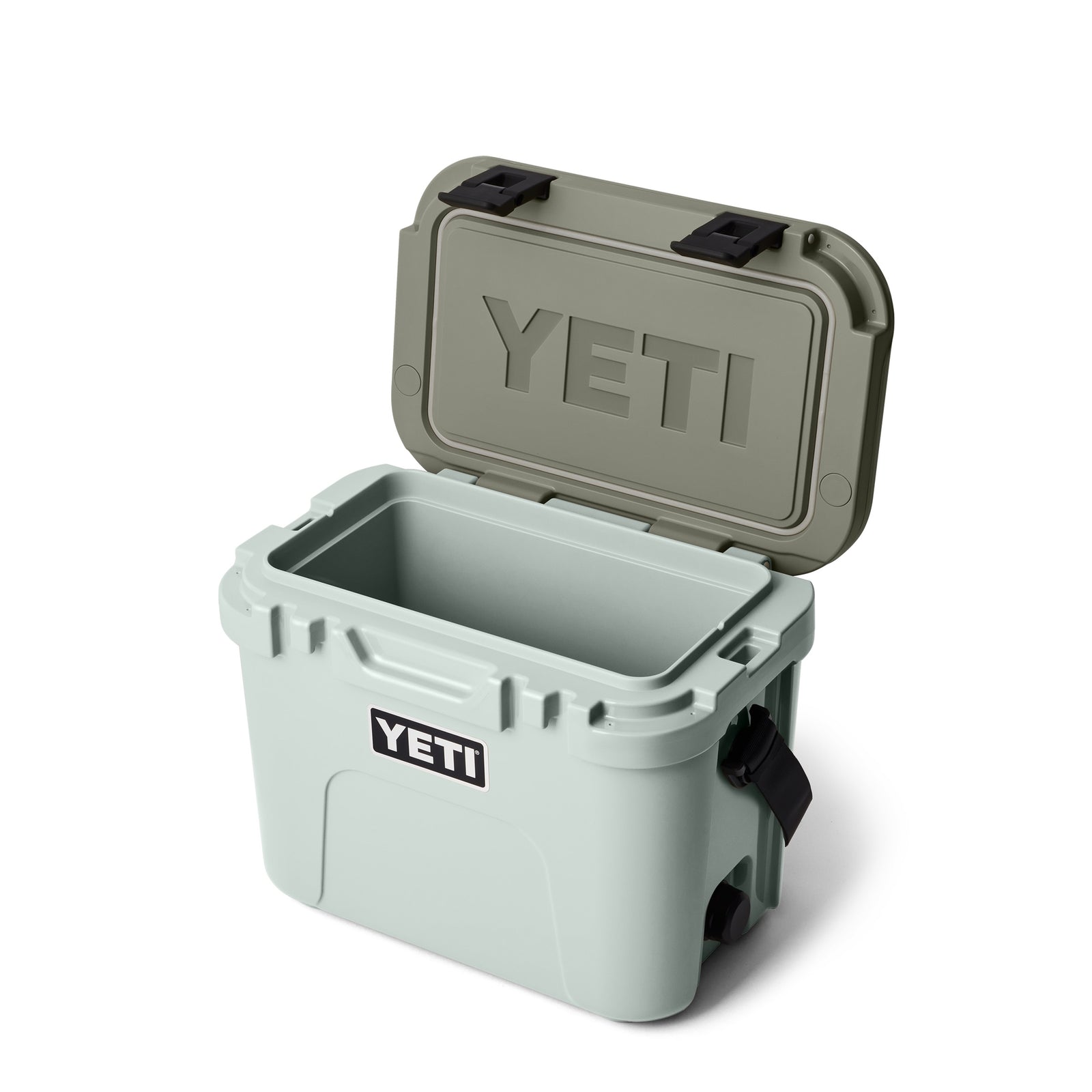 Eine hellgraue YETI® Kühlbox Roadie 15 - Ridgeline steht mit geöffnetem, olivgrünem Klappdeckel vor weißem Hintergrund und zeigt einen leeren Innenraum. Auf der Vorderseite ist das YETI-Logo zu sehen, ergänzt durch passende olivgrüne Verschlüsse.