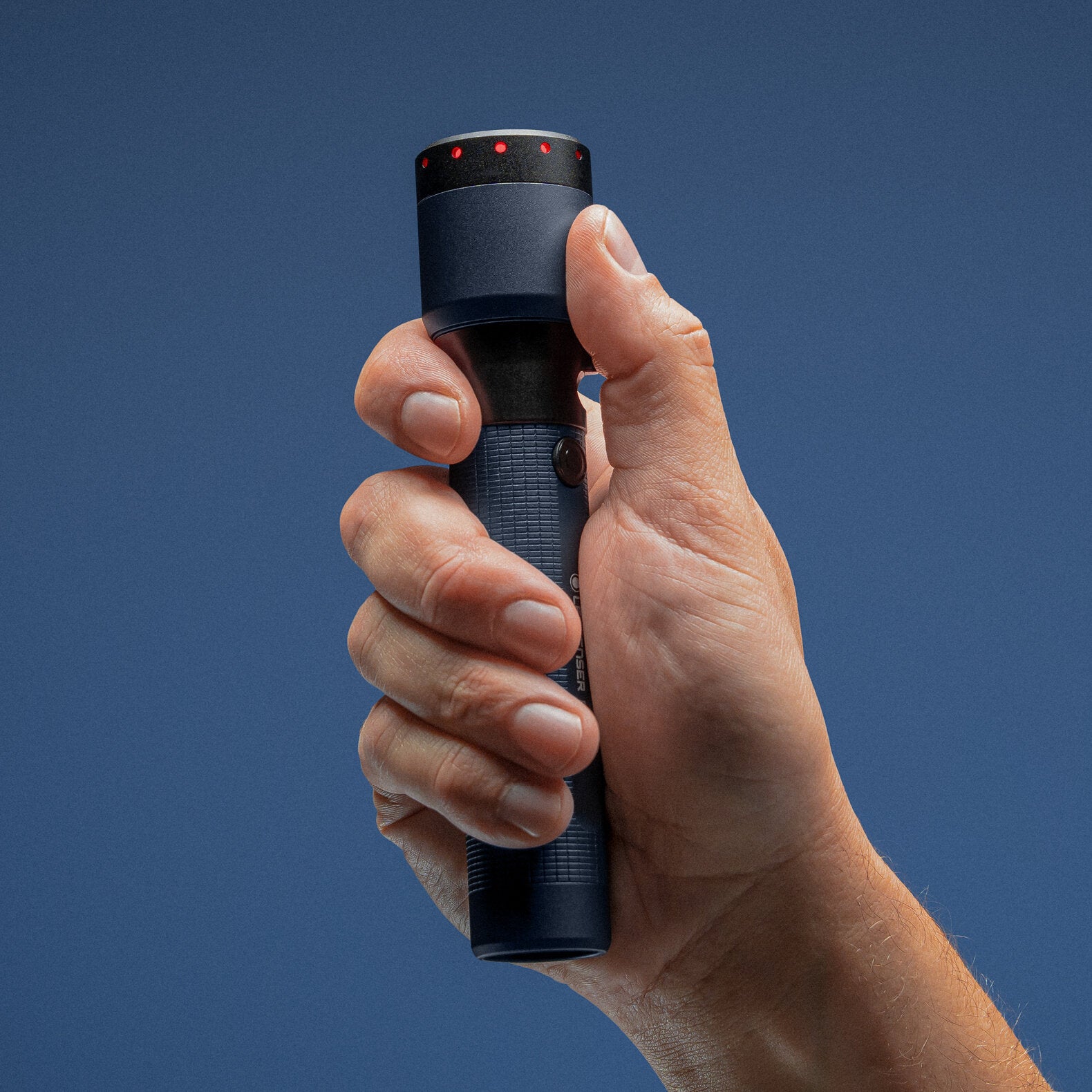 Eine Hand hält die LEDLENSER Taschenlampe P7R - 25 Jahre Edition aufrecht vor einem schlichten blauen Hintergrund. Diese Jubiläums-Taschenlampe verfügt über einen strukturierten Griff und rote Punkte um die Linse, die Stil mit Leistung kombinieren.