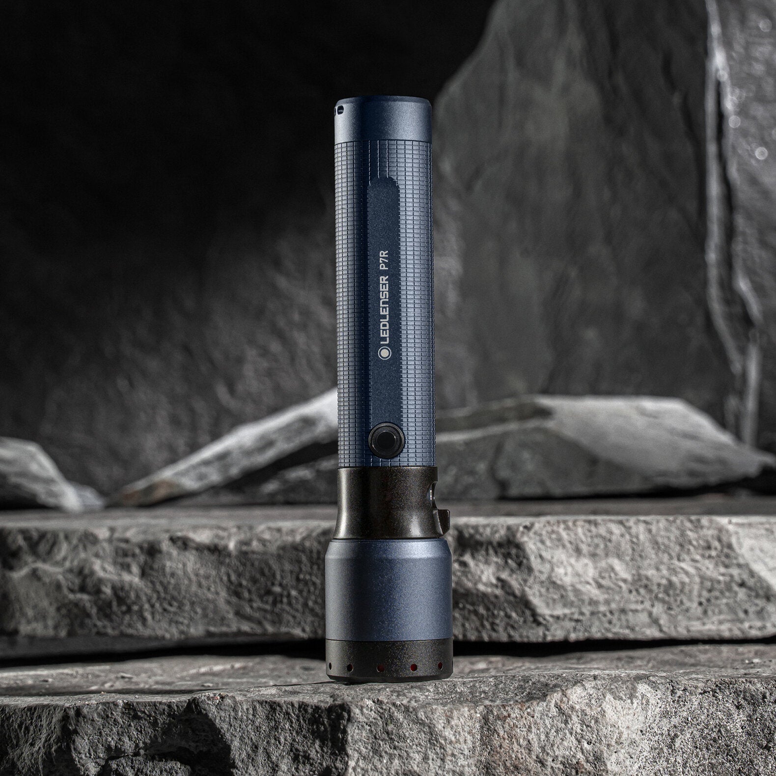 Die LEDLENSER Taschenlampe P7R - 25 Jahre Edition, in dunkelblau und schwarz, steht aufrecht auf gestapelten grauen Felsen. Diese besondere Taschenlampe hat einen strukturierten Griff, einen sichtbaren Knopf und liefert 2000 Lumen Helligkeit.