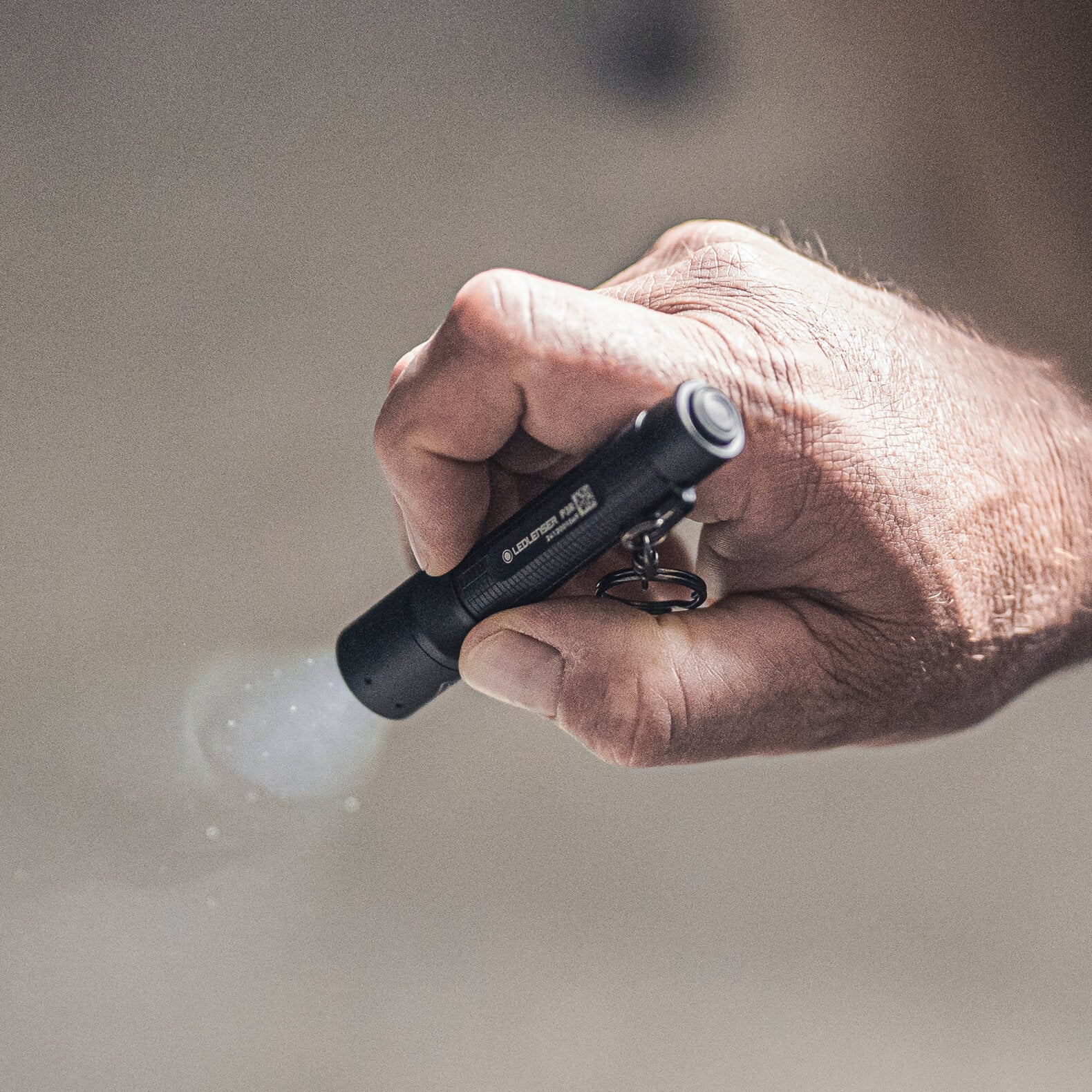 Nahaufnahme einer Person, die eine LEDLENSER Taschenlampe P3R in der Hand hält, die eingeschaltet ist und einen hellen Lichtstrahl in eine schwach beleuchtete Umgebung wirft.