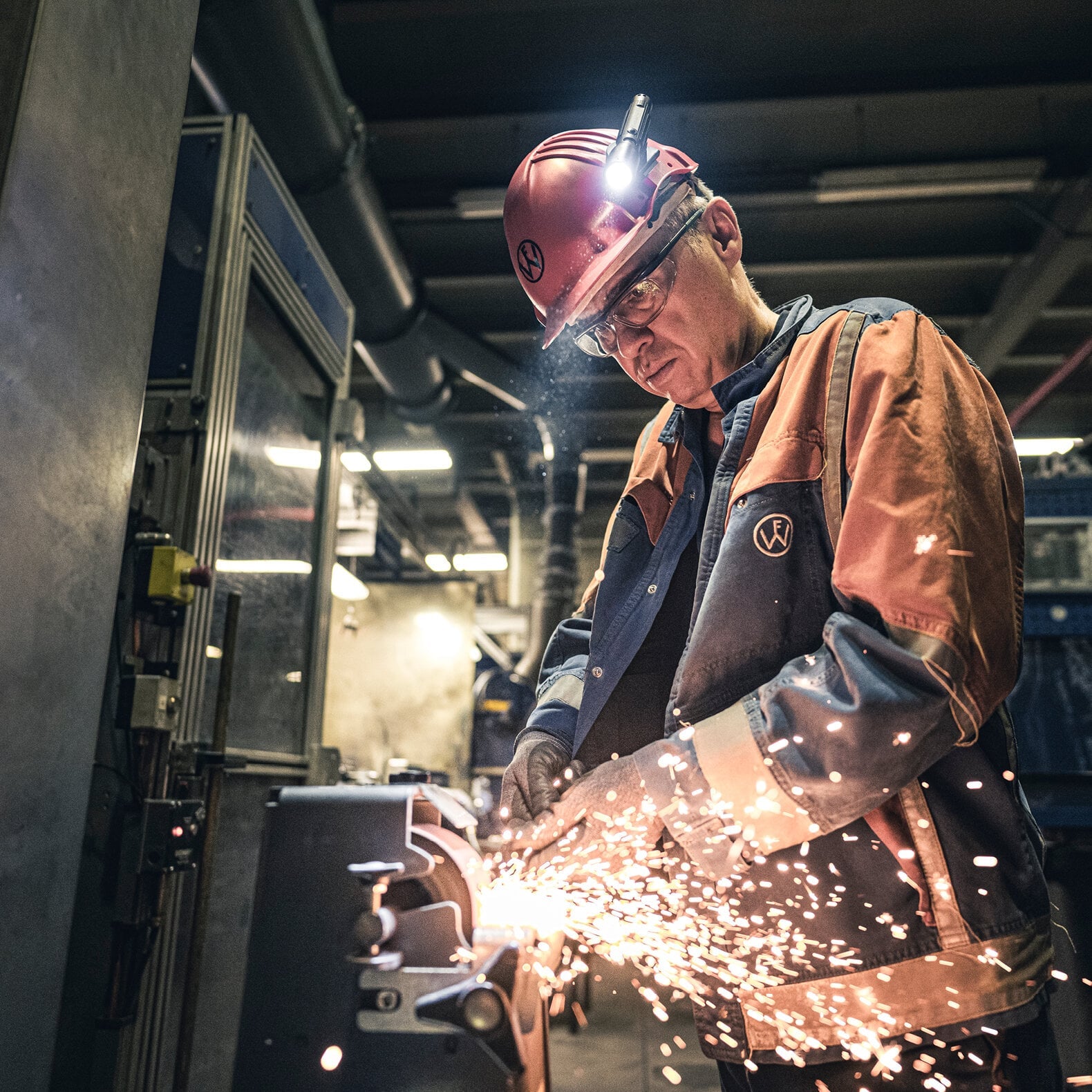 Ein Arbeiter in Schutzkleidung, Helm mit LEDLENSER Ledlenser Taschenlampe P5R mit Advanced Focus System, benutzt in einer Industriehalle ein Schleifwerkzeug, das helle Funken sprüht.