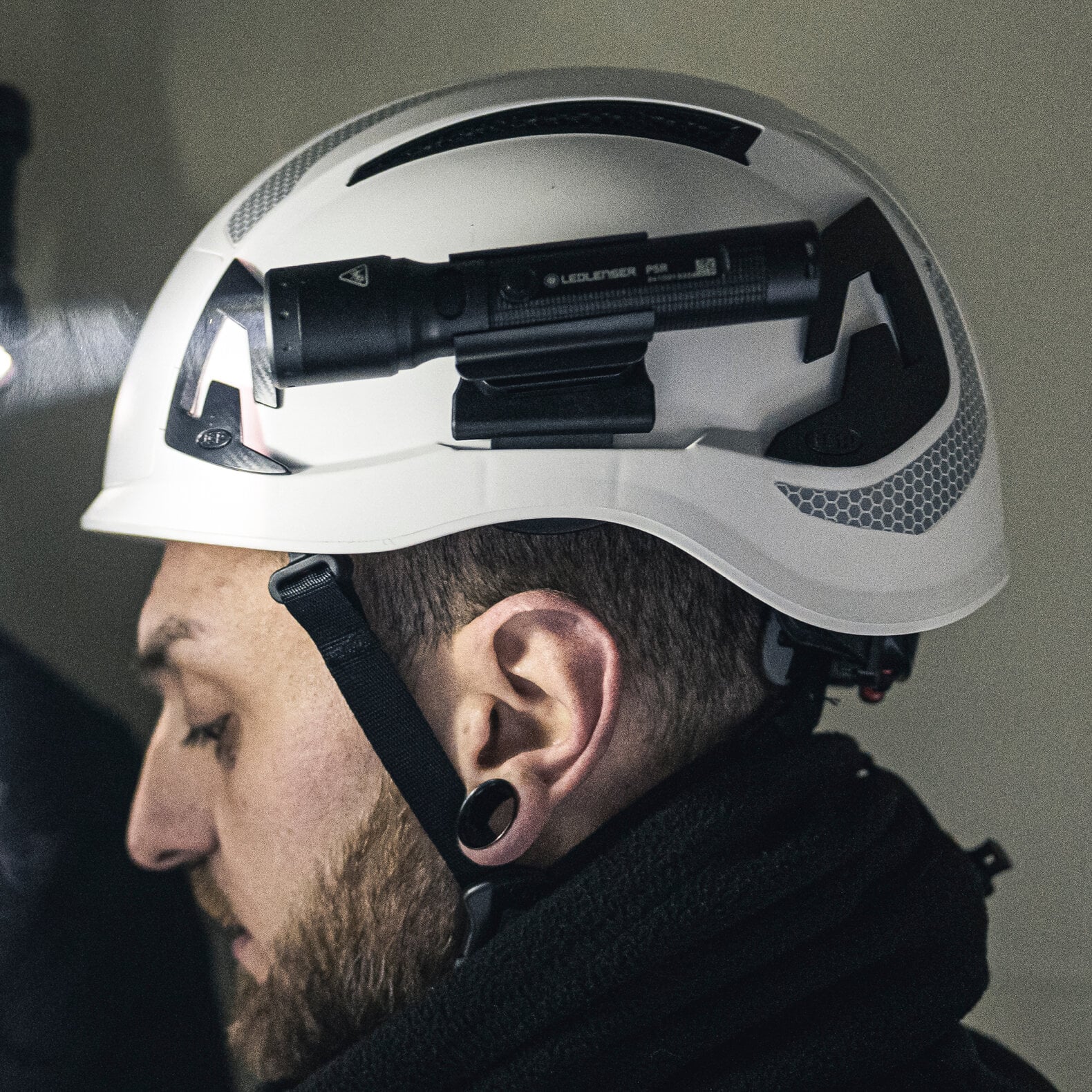 Ein Seitenprofil zeigt eine Person mit dunklem Bart, schwarzen Ohrenschützern und einem weißen Schutzhelm (mit Kinnriemen, schwarzen Akzenten), der mit einer LEDLENSER Ledlenser Taschenlampe P5R mit Advanced Focus System ausgestattet ist.