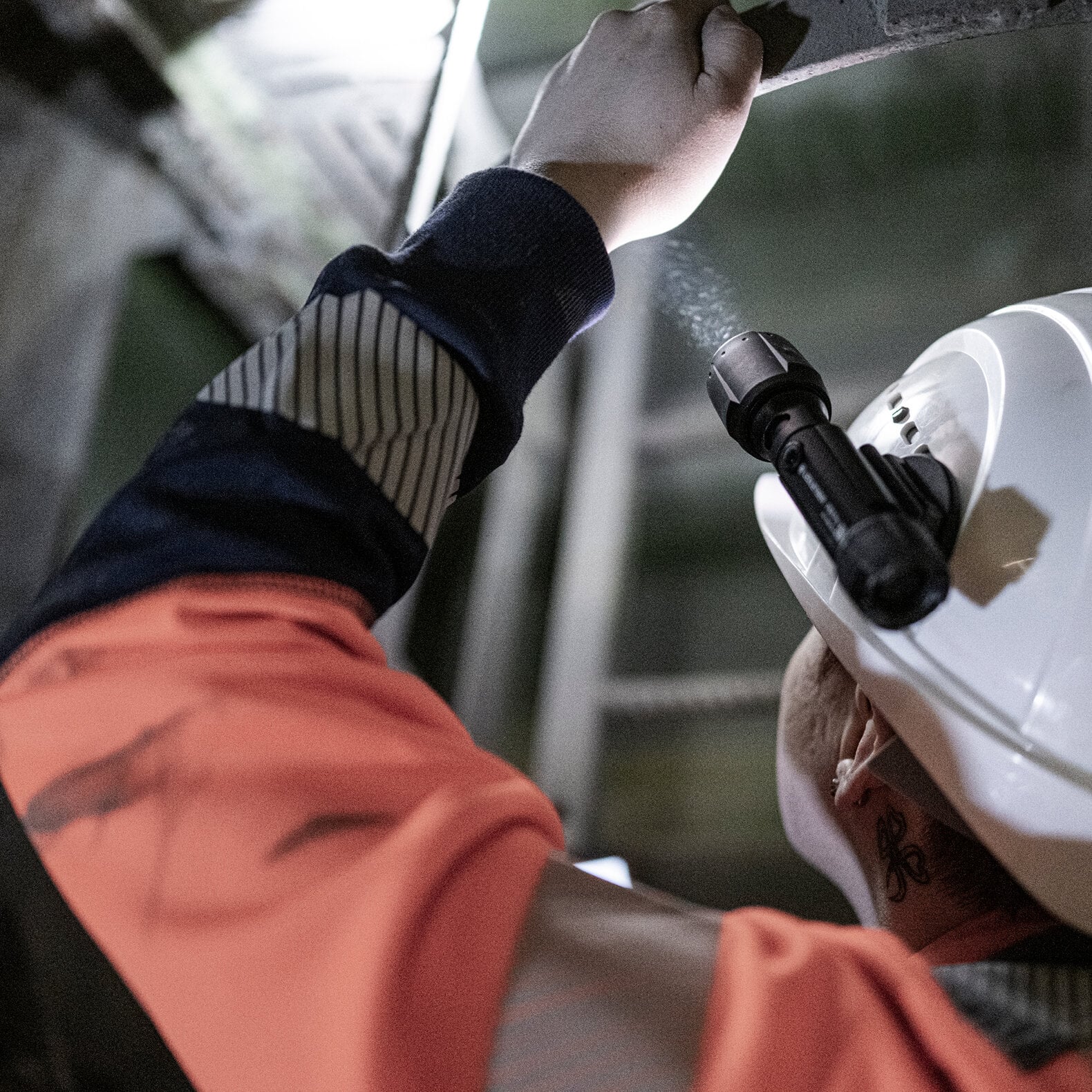 Eine Person, die eine orangefarbene Sicherheitsjacke und einen weißen Schutzhelm mit einer LEDLENSER Ledlenser Taschenlampe P7R Pro trägt, inspiziert eine Überkopfkonstruktion in einer industriellen Außenumgebung.