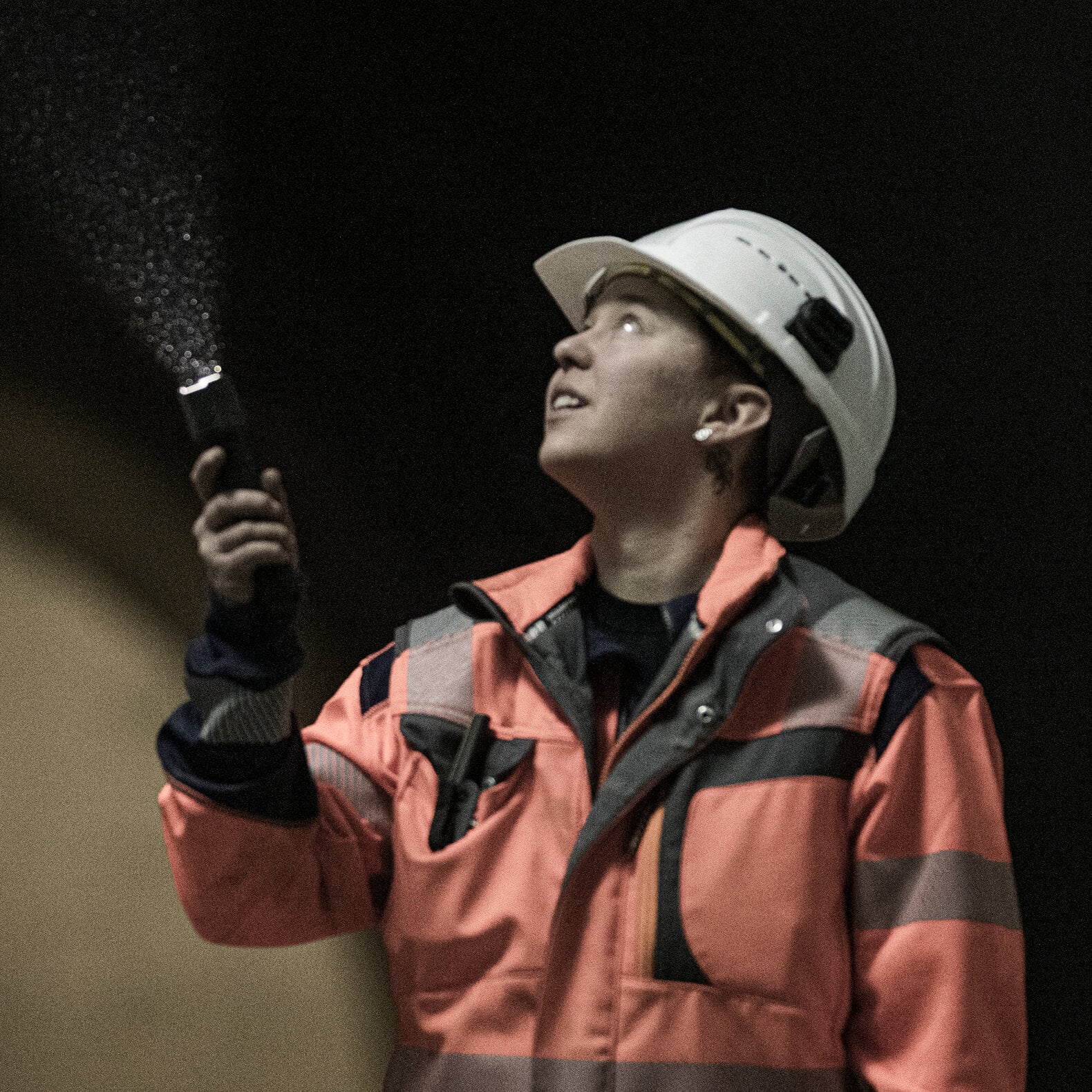 Eine Person mit weißem Schutzhelm und orangefarbener Warnweste richtet eine LEDLENSER Ledlenser Taschenlampe P7R Pro nach oben, um bei einer Inspektion im Freien feine Wassertropfen zu beleuchten, während sie in dieselbe Richtung schaut.