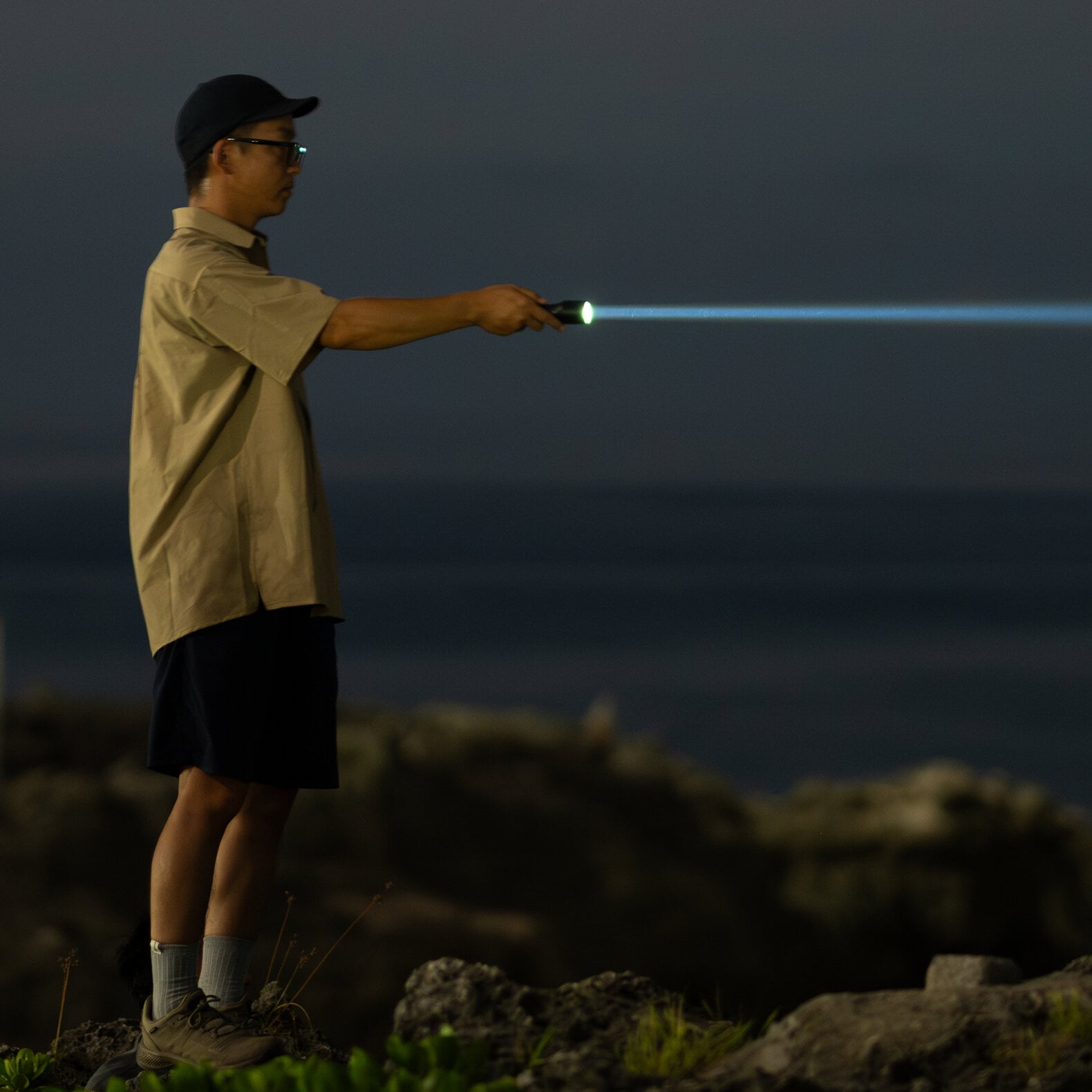 Eine Person mit Mütze, Brille, beigem Hemd und kurzen Hosen steht nachts auf felsigem Grund und leuchtet mit einer LEDLENSER Ledlenser Taschenlampe P9R Core LEP in die Ferne. Der dunkle Hintergrund lässt eine Andeutung von Wasser oder Himmel erkennen.