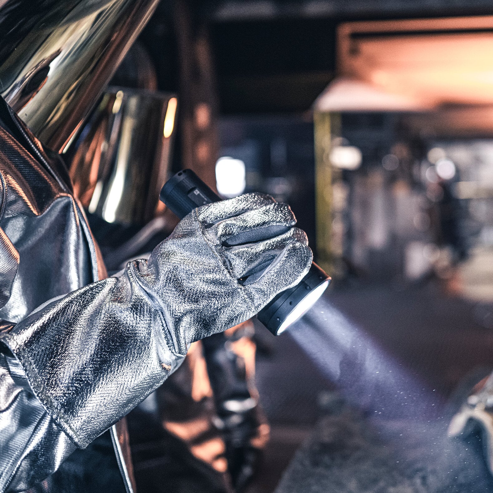 Bekleidet mit einem reflektierenden Metallanzug und Handschuhen leuchtet eine Person mit der LEDLENSER Taschenlampe P18R in einer Industrie- oder Werkstattumgebung eine Fläche mit ihrem starken 4500-Lumen-Strahl aus.