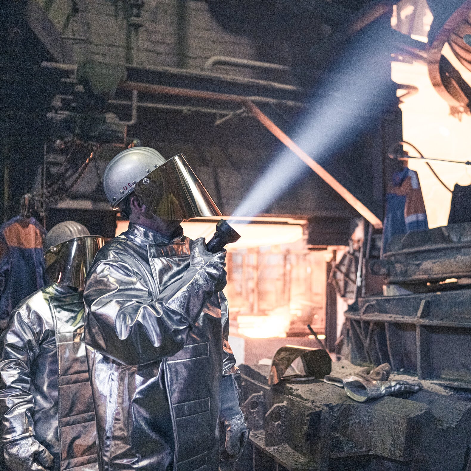 Arbeiter in reflektierenden Anzügen und Helmen verwenden eine LEDLENSER Taschenlampe P18R, um Gießereianlagen zu überprüfen, während im Hintergrund geschmolzenes Metall glüht.