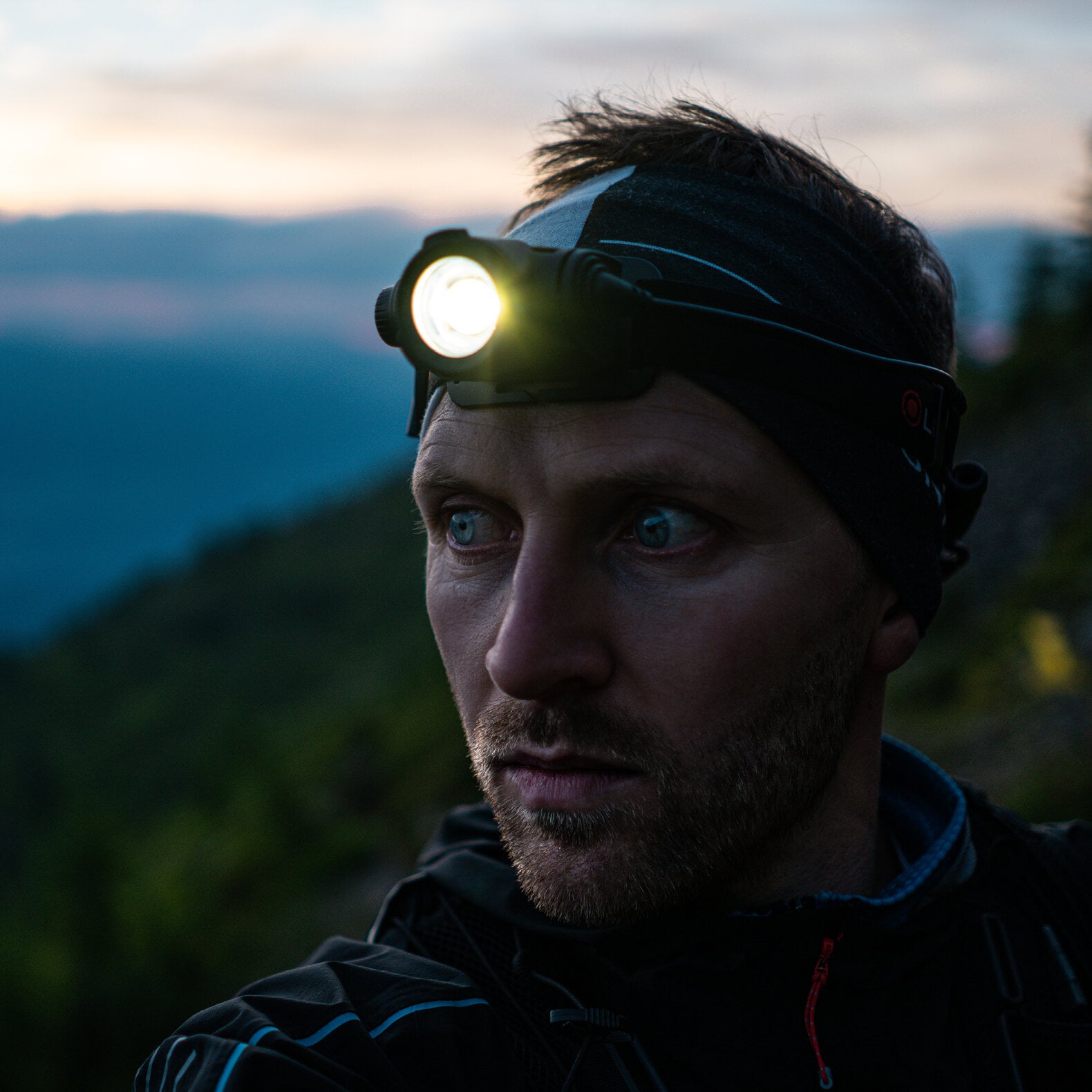 Ein Mann, der die LEDLENSER Stirnlampe H7R Core mit Advanced Focus System trägt, blickt in der Dämmerung aufmerksam zur Seite, Berge und Bäume verschwimmen sanft im Hintergrund.