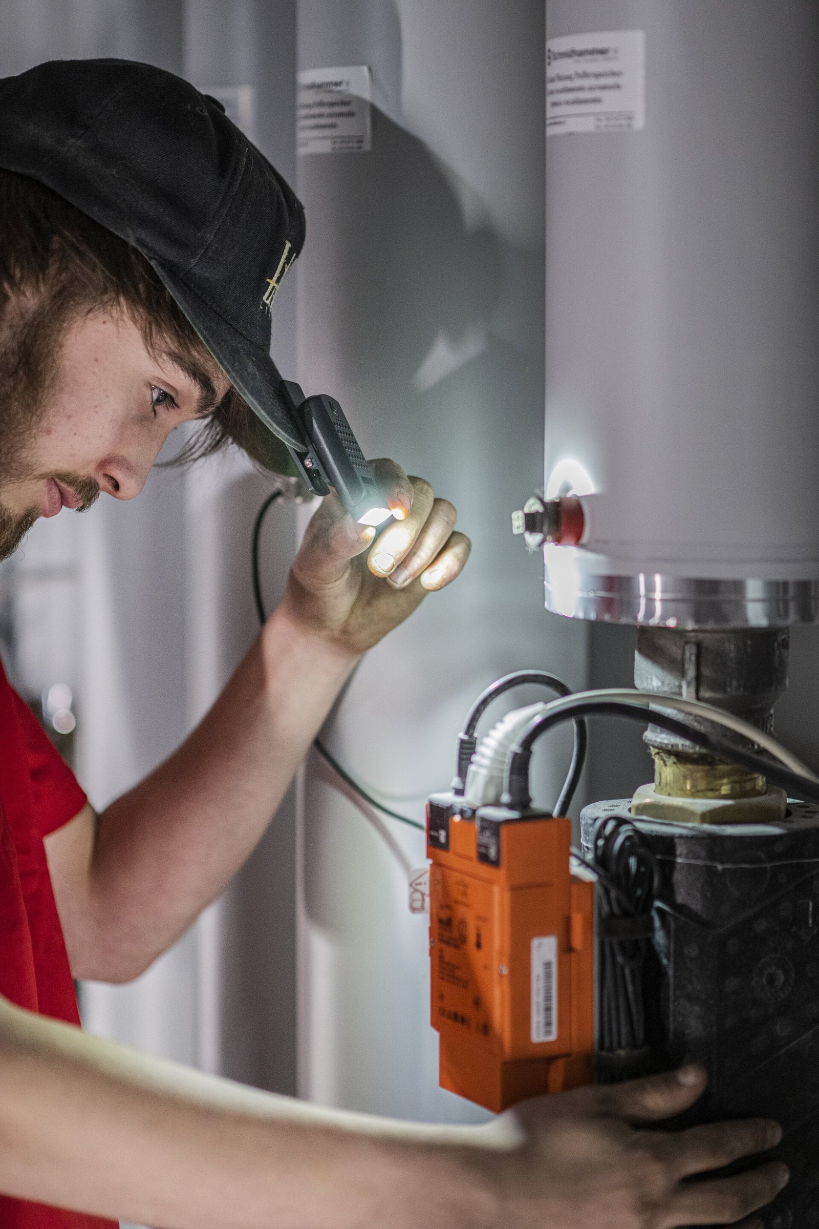 Eine Person mit Mütze und rotem Hemd verwendet die LEDLENSER Ledlenser W1R Work, um Kabel und Anschlüsse an Industrieanlagen in der Nähe eines großen grauen Zylinders zu beleuchten.