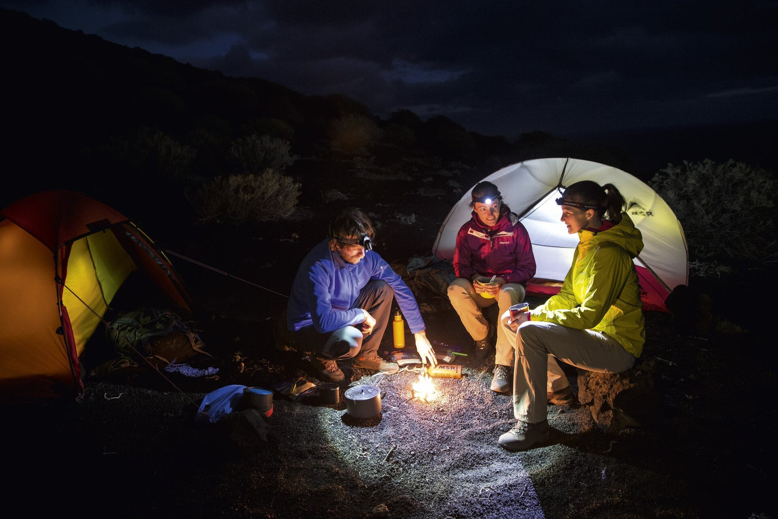 Drei Personen sitzen nachts an einem kleinen Lagerfeuer zwischen zwei Zelten, tragen Outdoor-Jacken und eine LEDLENSER Stirnlampe H14R.2 mit Advanced Focus System und Akku und teilen im sanften Schein warme Getränke und Speisen.
