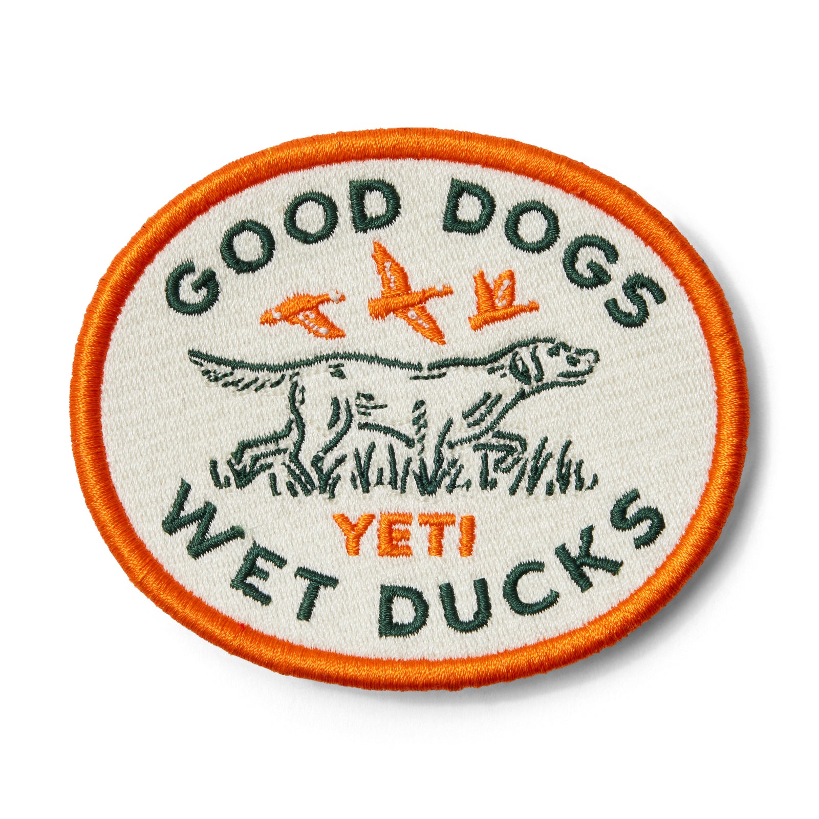 Das YETI® Good Dogs Patch von YETI zeigt eine Stickerei mit einem Hund, zwei Enten und dem Text "GOOD DOGS WET DUCKS YETI!" auf weißem Grund mit orangefarbenem Rand. Klettfläche kompatibel - perfekt für Outdoor-Ausrüstung.