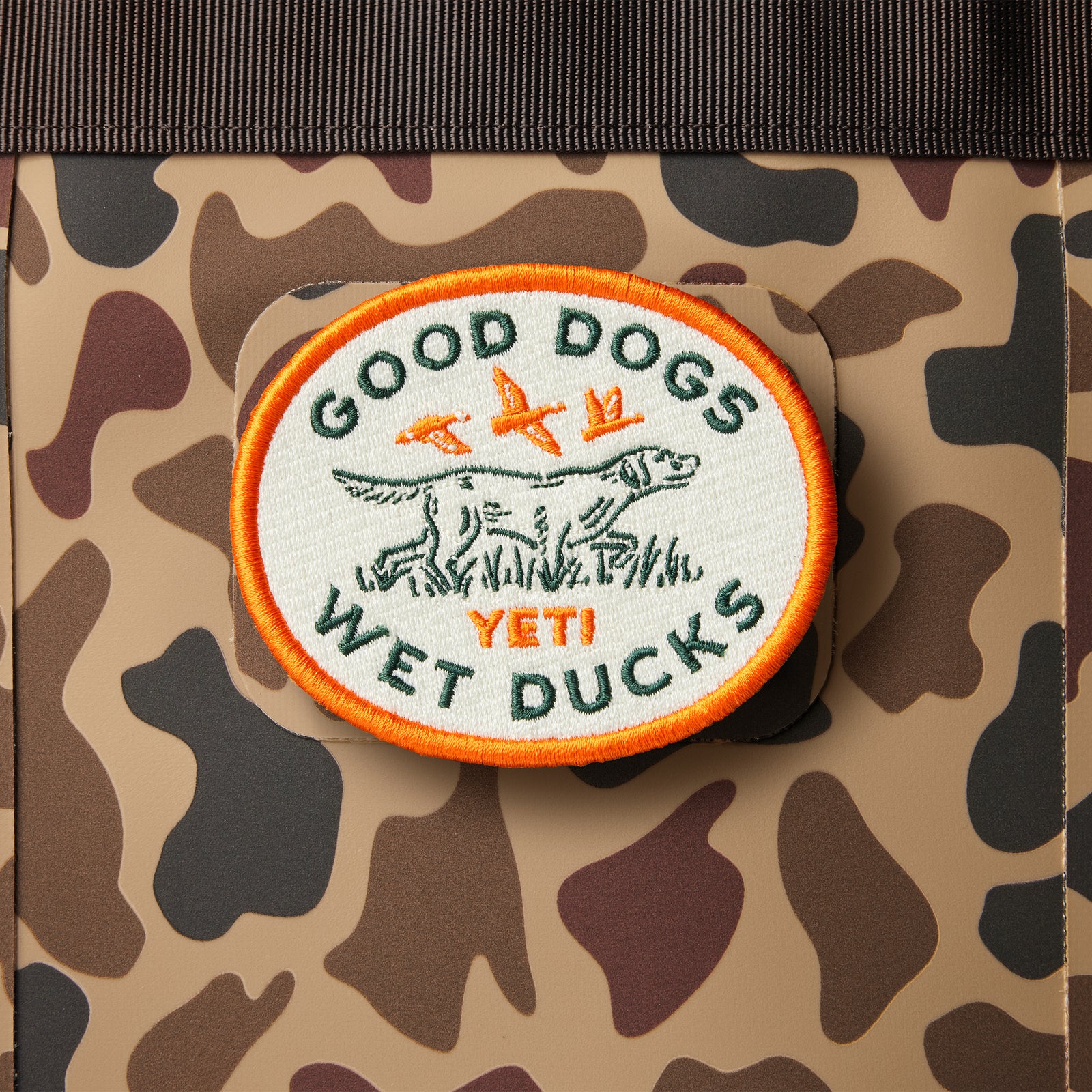 Das YETI® Good Dogs Patch von YETI zeigt einen Hund, zwei fliegende Enten und den Text "GOOD DOGS WET DUCKS YETI" auf einem Camouflage-Hintergrund mit orangefarbenem Rand; Klettfläche-kompatibel für Ihre Outdoor-Ausrüstung.