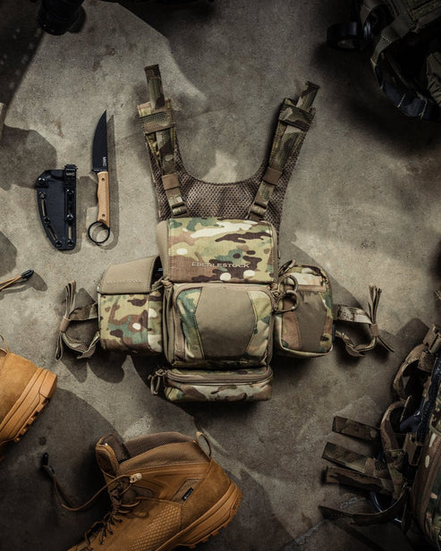 Taktische Tarnweste mit mehreren Taschen auf Betonboden, umgeben von braunen Kampfstiefeln, einem schwarzen Klappmesser, einem Messer mit feststehender Klinge und EBERLESTOCK Recon Modular Bino Pack Large Jagd Ausrüstung.