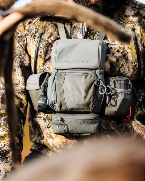 Eine Person in Tarnjacke und einem EBERLESTOCK Recon Modular Bino Pack Large - einem fortschrittlichen Ausrüstungsträger für den Outdoor-Einsatz - wird im Freien gezeigt, mit unscharfen Ästen im Vordergrund.