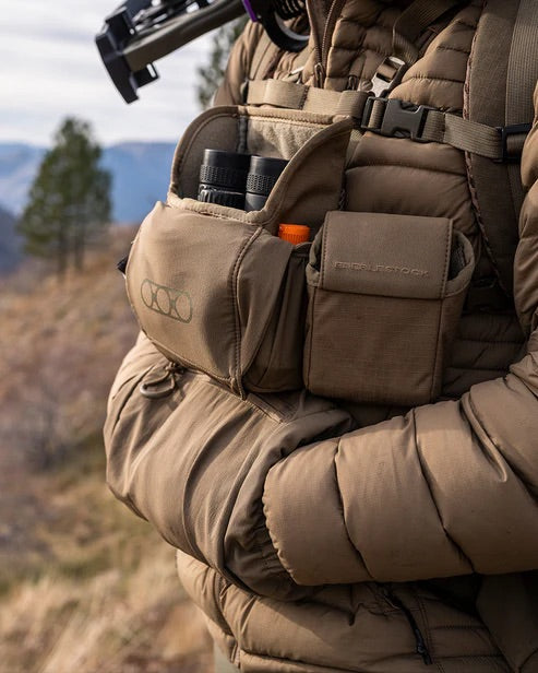 Eine Person im Freien, mit brauner Pufferjacke und Rucksack, hält das EBERLESTOCK Recon Modular Bino Pack Large, gefüllt mit einem Fernglas - ein praktisches Tragesystem - und einem kleinen orangen Gegenstand. Bäume und Berge erscheinen verschwommen hinter ihnen.