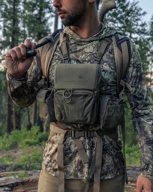 Ein Mann in Tarnkleidung und einer taktischen Weste steht in einem Wald und trägt sein EBERLESTOCK Recon Modular Bino Pack Large, das vor dem Hintergrund von Bäumen und Grünflächen Jagdausrüstung der Spitzenklasse zeigt.