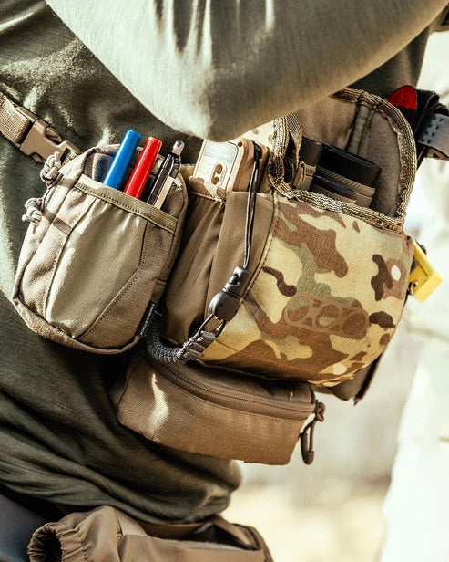 Nahaufnahme einer Person in einem grünen, langärmeligen Hemd, die einen EBERLESTOCK Recon Modular Bino Pack Large mit mehreren Tarntaschen trägt, in denen Stifte, Marker, ein Telefon und kleinere Ausrüstungsgegenstände untergebracht sind.