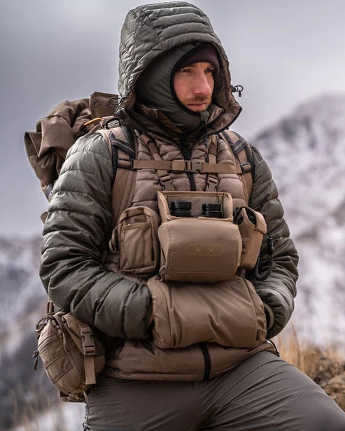Ausgestattet mit schwerer Outdoor-Ausrüstung und Kapuze steht eine Person mit Rucksack und dem EBERLESTOCK Eberlestock Recon Modular Bino Pack Large vor verschneiten Bergen und Wolken - eine Top-Wahl für jede Jagdausrüstungstour.