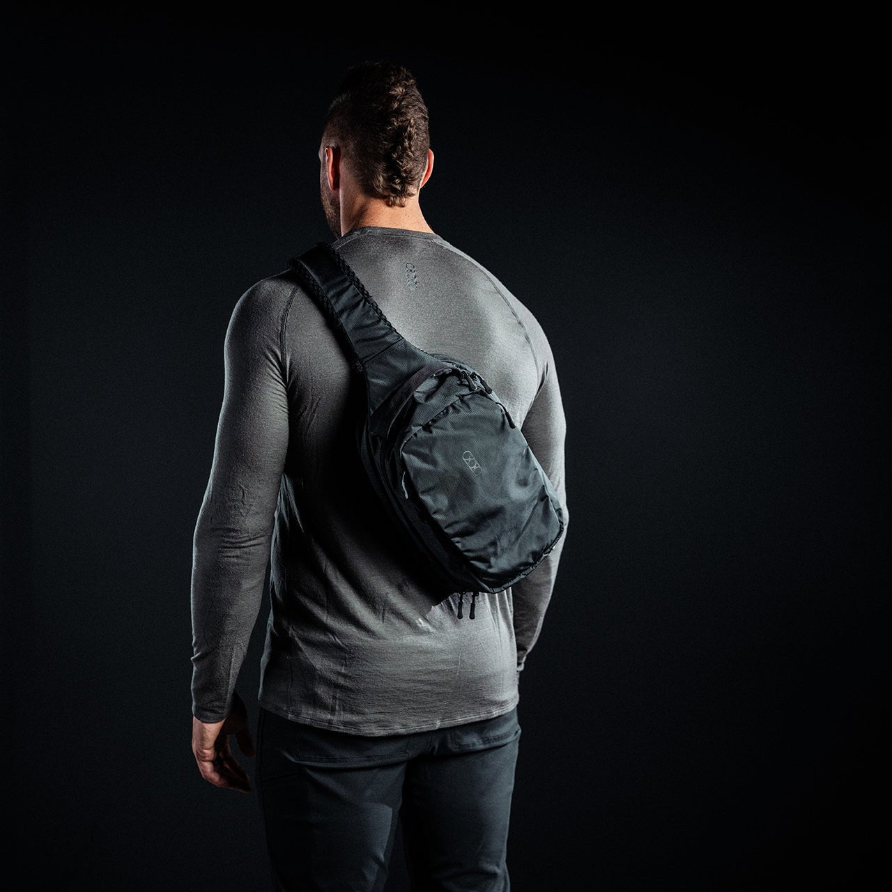 Ein Mann in einem grauen, langärmeligen Hemd und einer schwarzen Hose steht mit dem Rücken zur Kamera und trägt einen EBERLESTOCK Fade Sling über der rechten Schulter - ideal zum diskreten Tragen einer Pistole - vor einem dunklen Hintergrund.