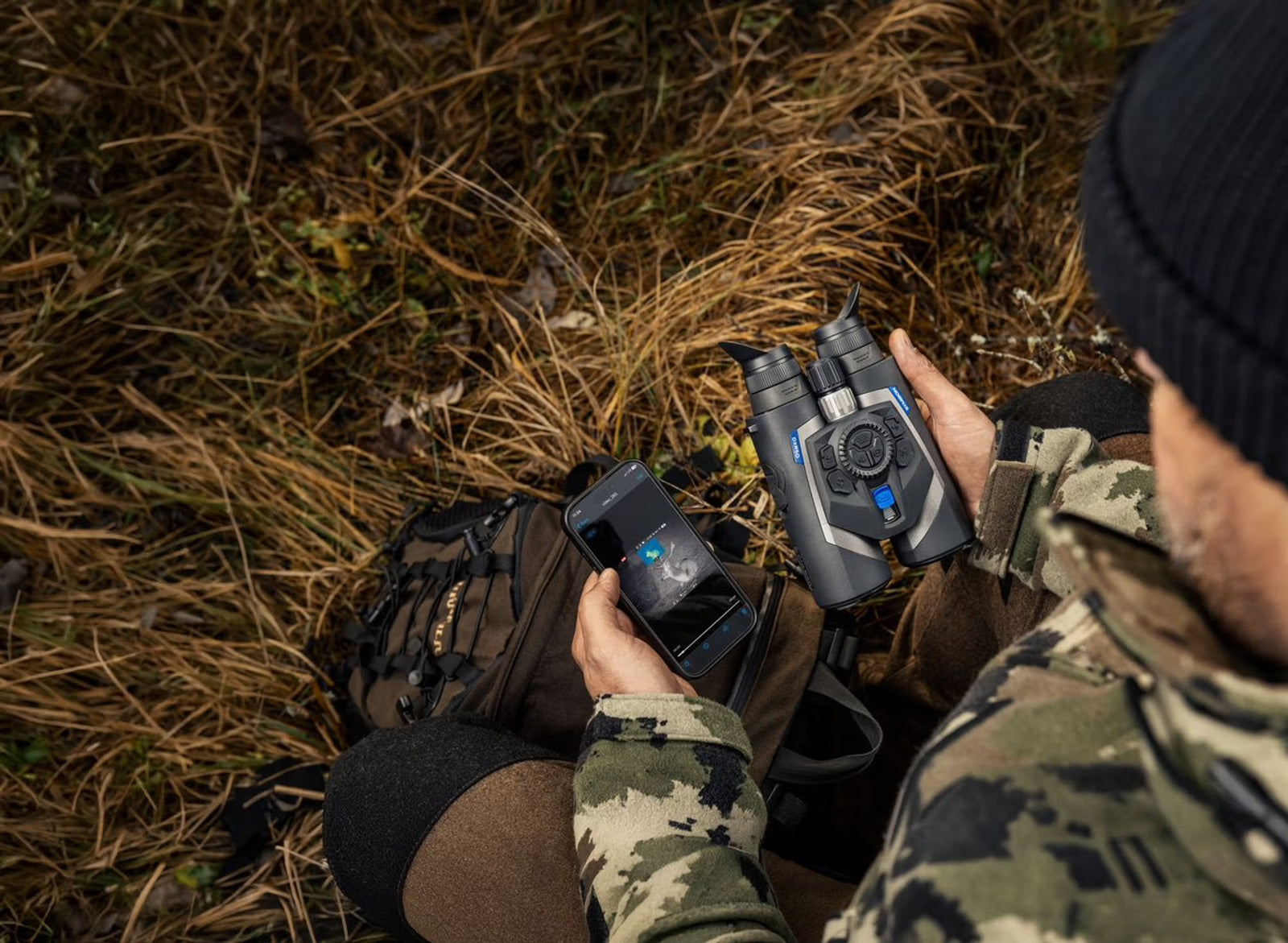 Eine getarnte Person sitzt im trockenen Gras, hält ein PULSAR Pulsar Multispektrales Fernglas Symbion LRF DXR50 in der einen und ein Smartphone mit Karten-App in der anderen Hand, einen Rucksack daneben.