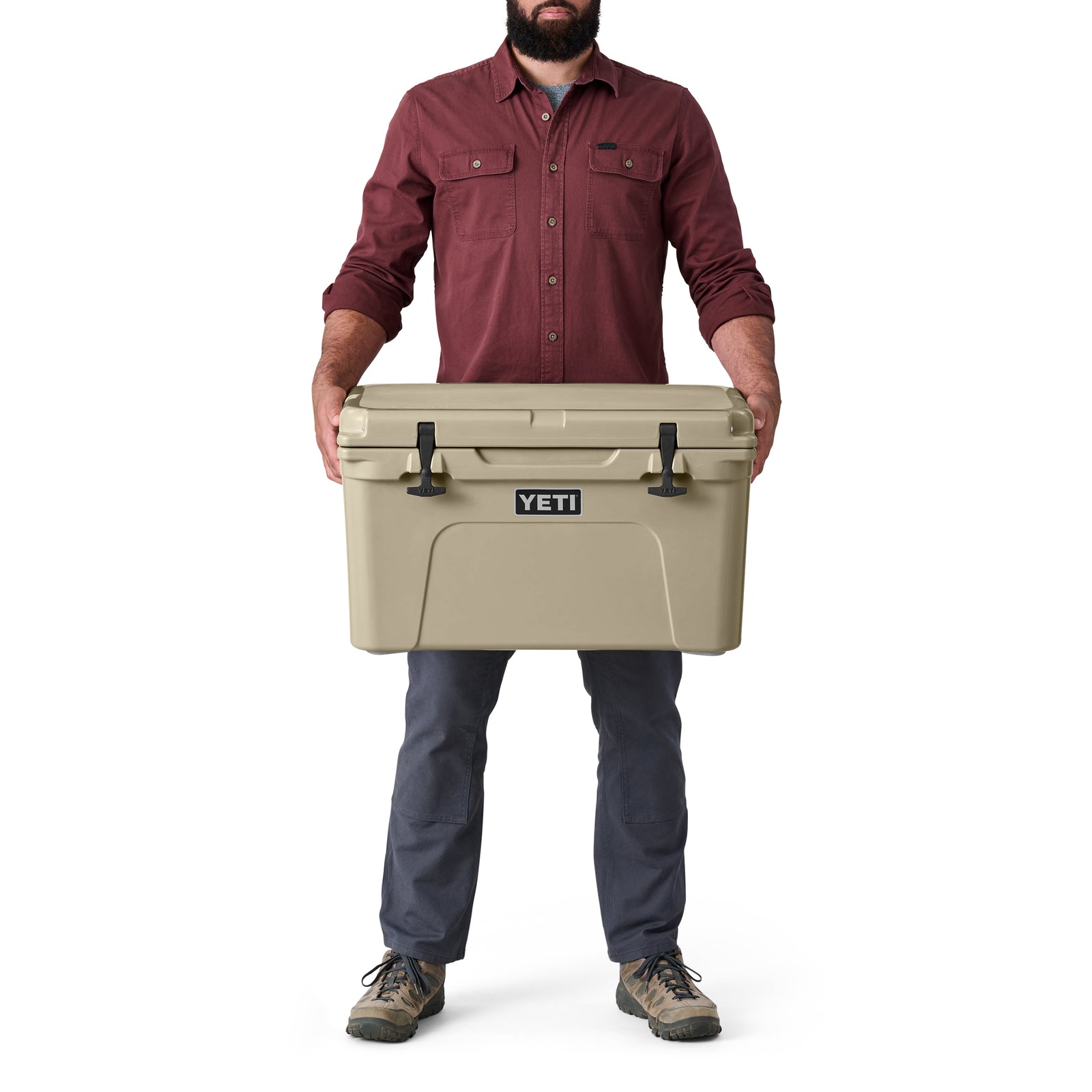 Ein bärtiger Mann in einem kastanienbraunen Hemd, einer blauen Hose und Wanderschuhen steht vor einem weißen Hintergrund und hält eine hellbraune YETI® Kühlbox Tundra 45 von YETI.