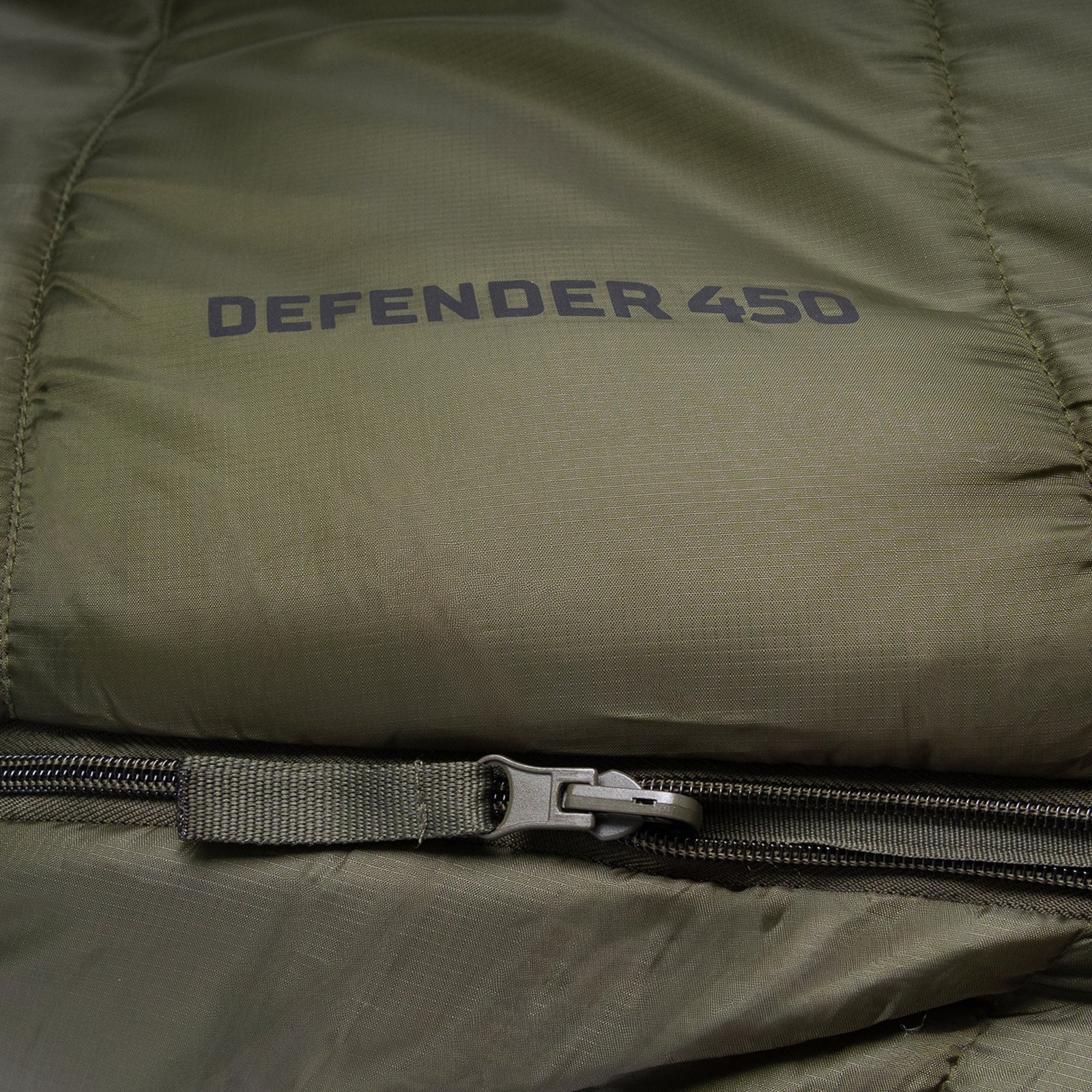 Nahaufnahme eines olivgrünen Highlander Schlafsacks Defender 450 von HIGHLANDER OUTDOOR, mit sichtbarem Reißverschluss und schwarzem Aufdruck "DEFENDER 450" auf dem Stoff.