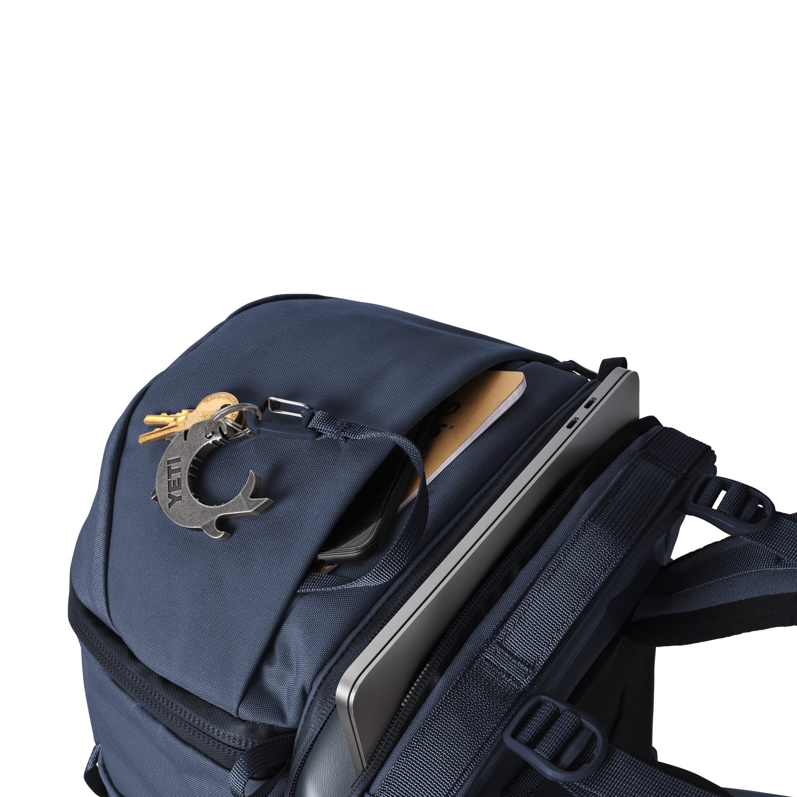 Der YETI® Rucksack Ranchero™ 27L in Classic Navy aus TuffSkin-Nylon hat ein offenes Fach, in dem ein Tablet und ein hellbraunes Notizbuch Platz finden, sowie Schlüssel mit einem fischförmigen Schlüsselanhänger am Gurt.