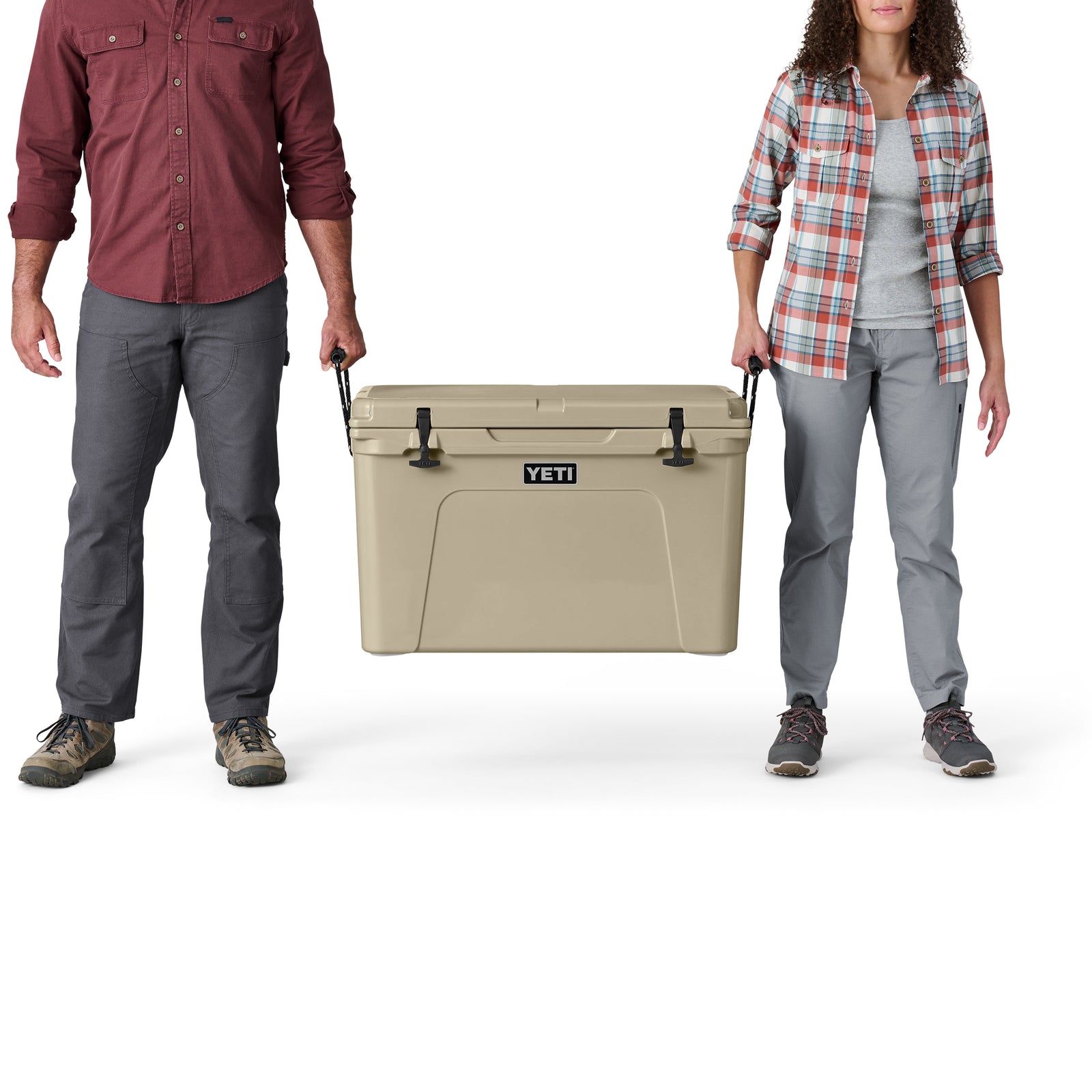 Ein Mann und eine Frau, beide in legerer Outdoor-Kleidung und Turnschuhen, stehen nebeneinander und halten eine große YETI®-Kühlbox „Tundra 105“ in der Farbe „Tan“ an ihren stabilen Seitengriffen fest – bereit für ihr nächstes Abenteuer.