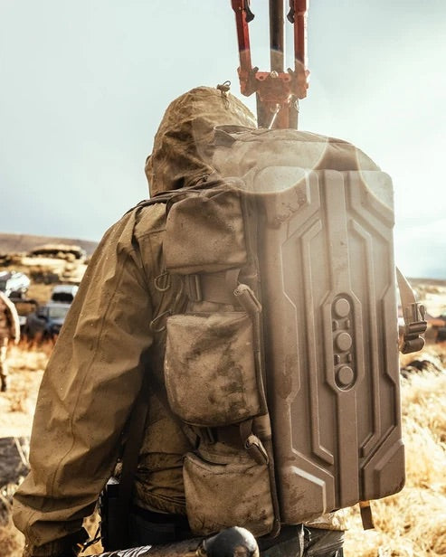 Eine Person in einer hellbraunen Kapuzenjacke steht im Freien auf einer trockenen, grasbewachsenen Fläche, den Rücken zur Kamera gewandt, im Hintergrund sind Sonnenlicht und Linsenreflexe zu sehen; sie trägt einen großen, robusten EBERLESTOCK UpRanger™-Rucksack.
