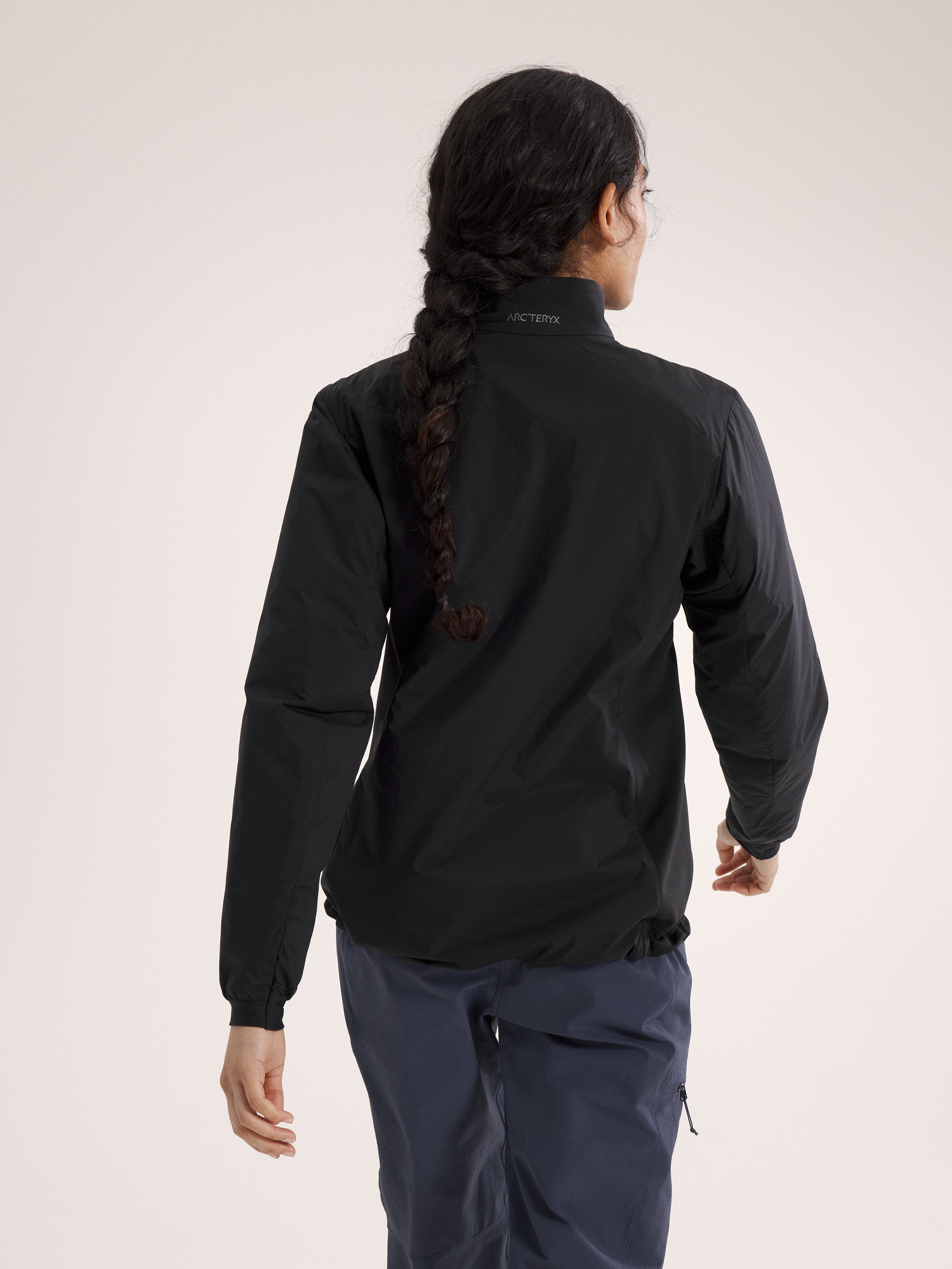 Eine Person mit langem, dunklem, geflochtenem Haar wird von hinten gezeigt, die eine blaue Hose und eine schwarze ARC'TERYX Atom Jacket Women's trägt und vor einem schlichten hellen Hintergrund steht.