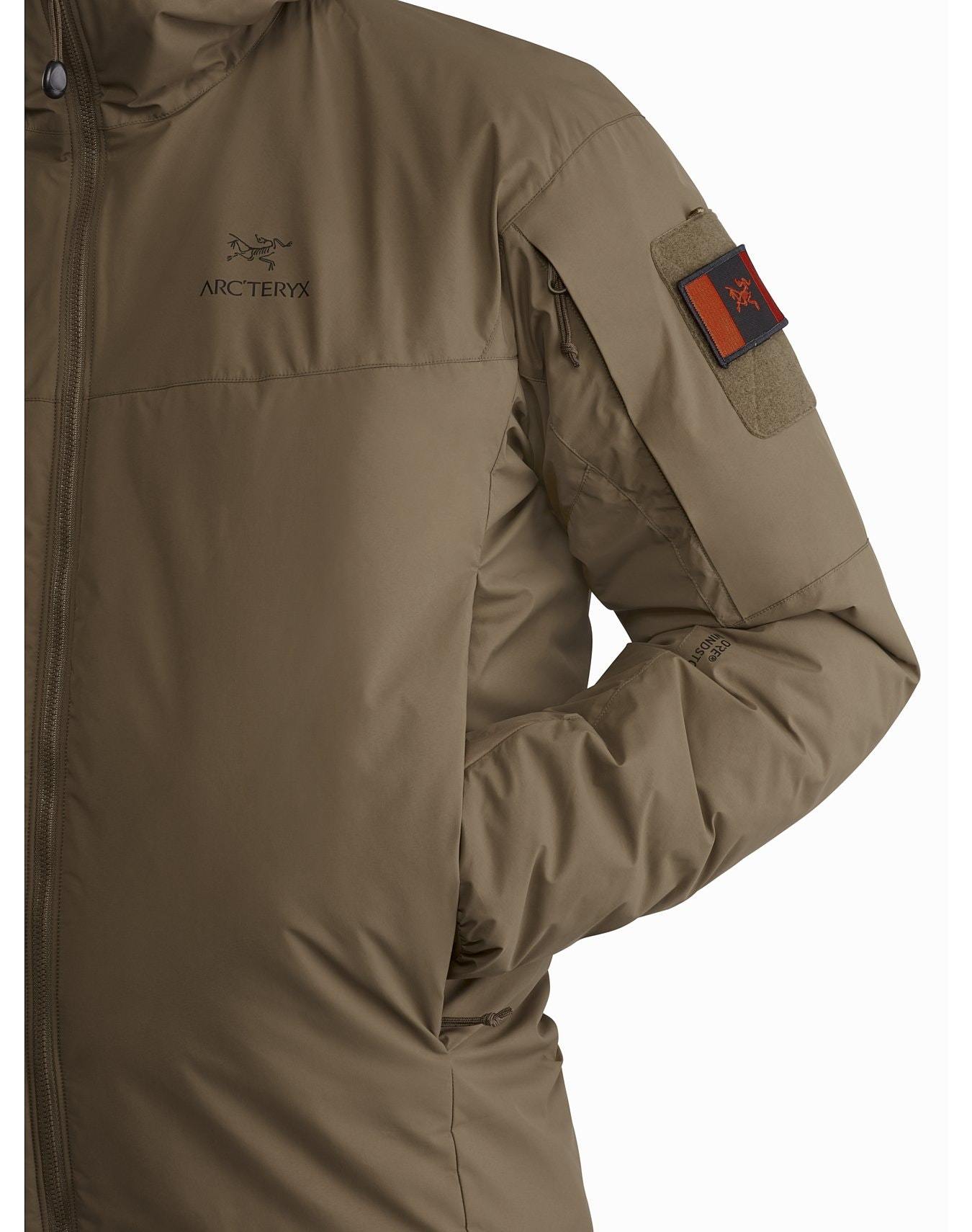 Nahaufnahme einer Person, die einen schwarzen ARC'TERYX LEAF Cold WX Hoody LT Gen2.1 trägt, mit dem Arc'teryx-Logo auf der Brust und einem kleinen roten Aufnäher auf der oberen Ärmeltasche. Der Kapuzenpullover verfügt über eine fortschrittliche CLIMASHIELD Isolierung für zusätzliche Wärme.