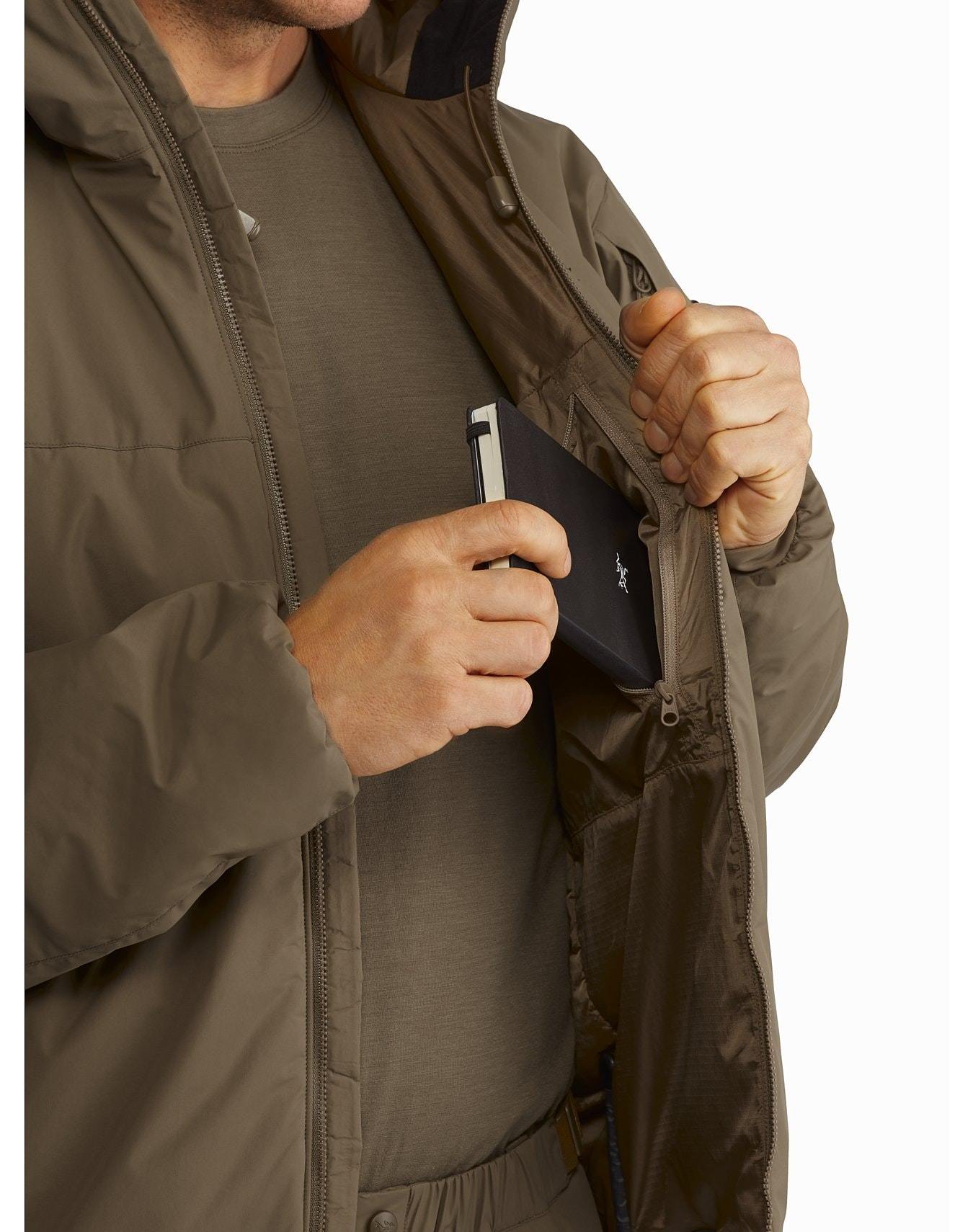 Eine Person, die den ARC'TERYX LEAF Cold WX Hoody LT Gen2.1 - Black trägt, steckt ein schwarzes Portemonnaie oder ein Handy in die innere Brusttasche ihrer Jacke, die sie offen hält, um den GORE WINDSTOPPER Schutz zu zeigen.