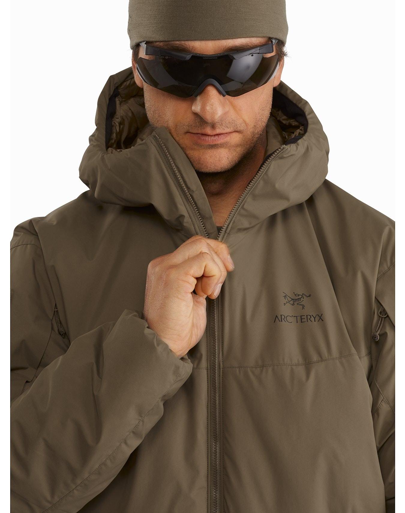 Eine Person, die einen rangergrünen ARC'TERYX LEAF Cold WX Hoody LT Gen2.1, eine dunkle Sonnenbrille und eine braune Mütze trägt, schließt mit neutraler Miene den Reißverschluss der Jacke.