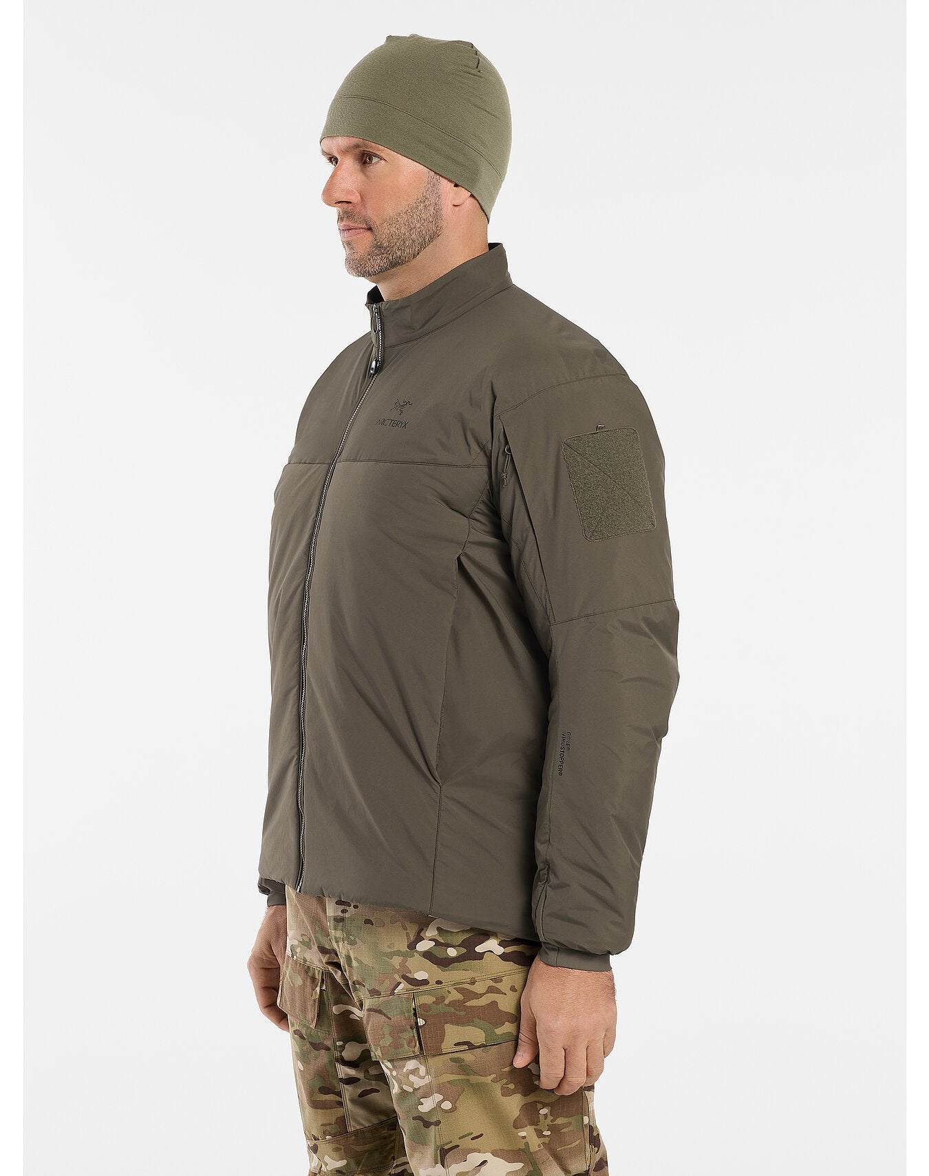 Ein Mann mit einer grünen Mütze, einer Tarnhose und einer ARC'TERYX LEAF Cold WX Jacket LT Gen2.1 (Black) mit einem Aufnäher auf dem Oberarm steht seitlich vor einem einfarbig weißen Hintergrund.