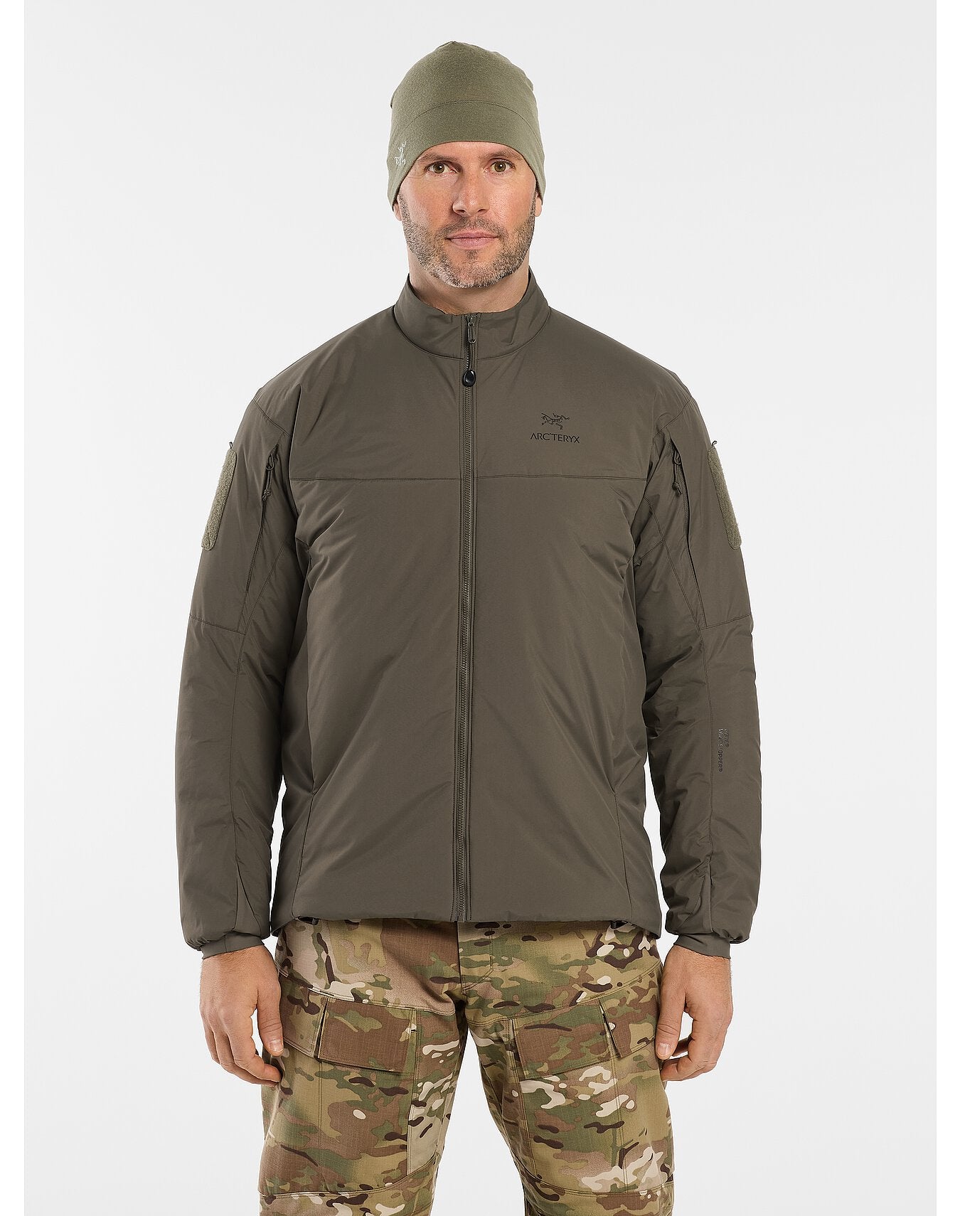 Ein Mann in einer ARC'TERYX LEAF Arc'teryx Cold WX Jacket LT Gen2.1 - Ranger Green und passender Mütze steht mit Blick nach vorne und trägt eine Tarnhose vor einem einfarbig weißen Hintergrund.