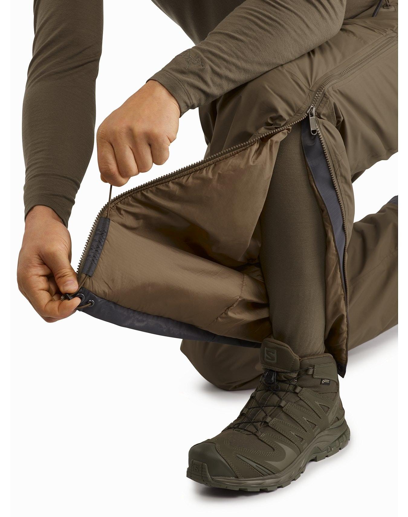 Eine Person kniet und öffnet den Reißverschluss an der Seite der schwarzen Arc'teryx LEAF Cold WX Pant LT Gen2 Hose, so dass eine Schicht darunter zum Vorschein kommt. Sie trägt dazu passende schwarze Wanderschuhe und ein langärmeliges Hemd.