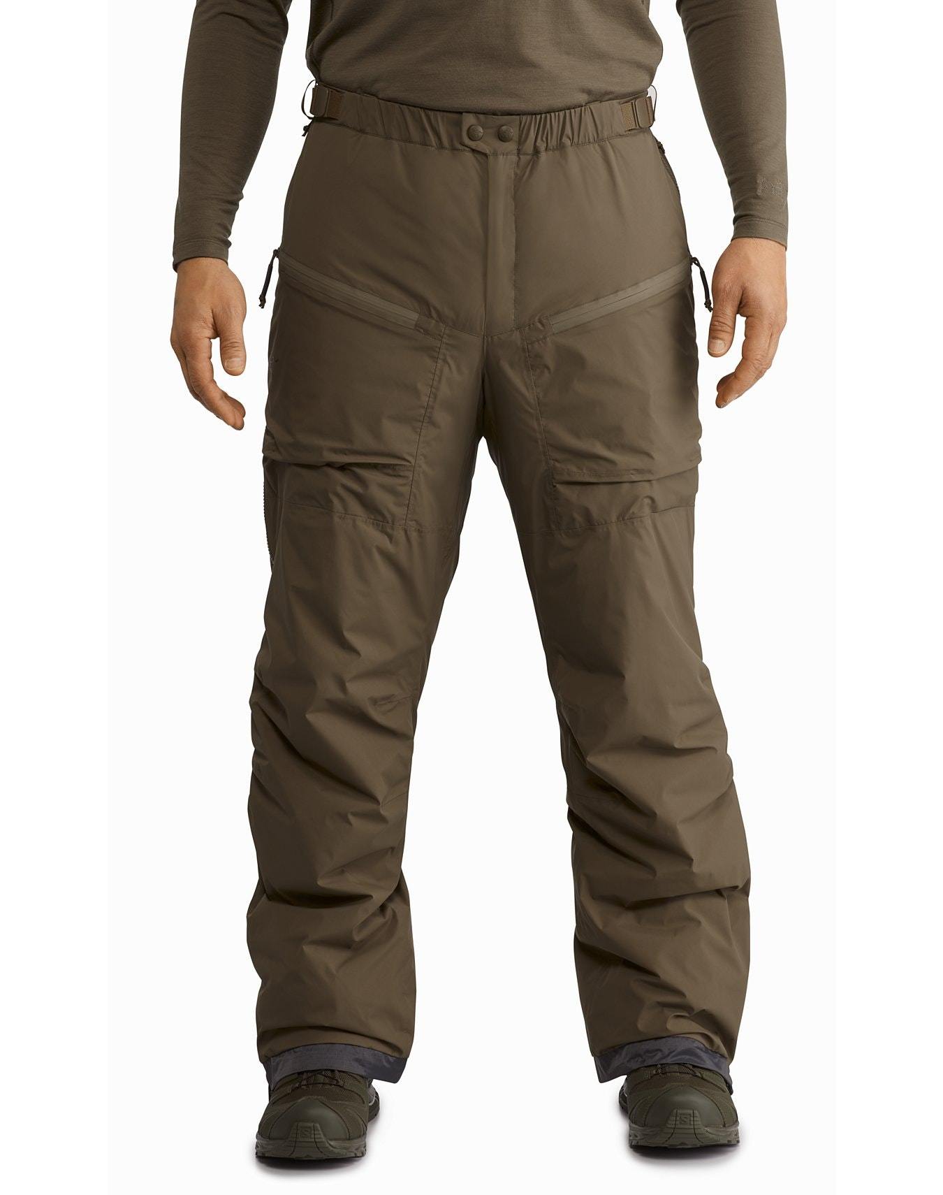 In der ARC'TERYX LEAF Arc'teryx Cold WX Pant LT Gen2 - Crocodile, einer isolierten Hose mit Reißverschlusstaschen, steht eine Person mit leicht gespreizten Beinen, gepaart mit einem langärmeligen Hemd und Wanderschuhen.