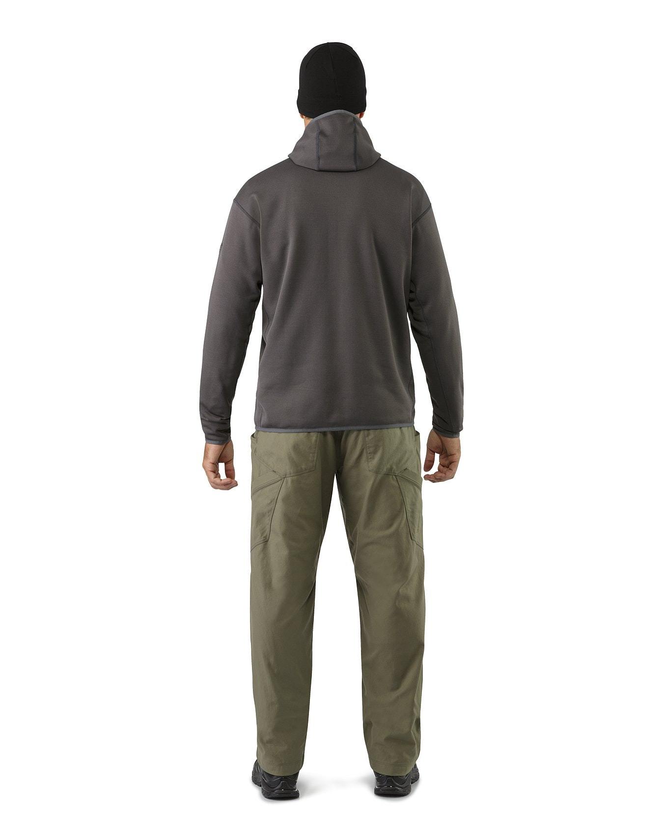 Eine Person blickt von der Kamera weg und trägt eine schwarze Mütze, den ARC'TERYX LEAF Naga Hoody Full Zip Gen2.1 - Crocodile als taktischen Midlayer, eine olivfarbene Cargohose und schwarze Schuhe vor einem weißen Hintergrund.