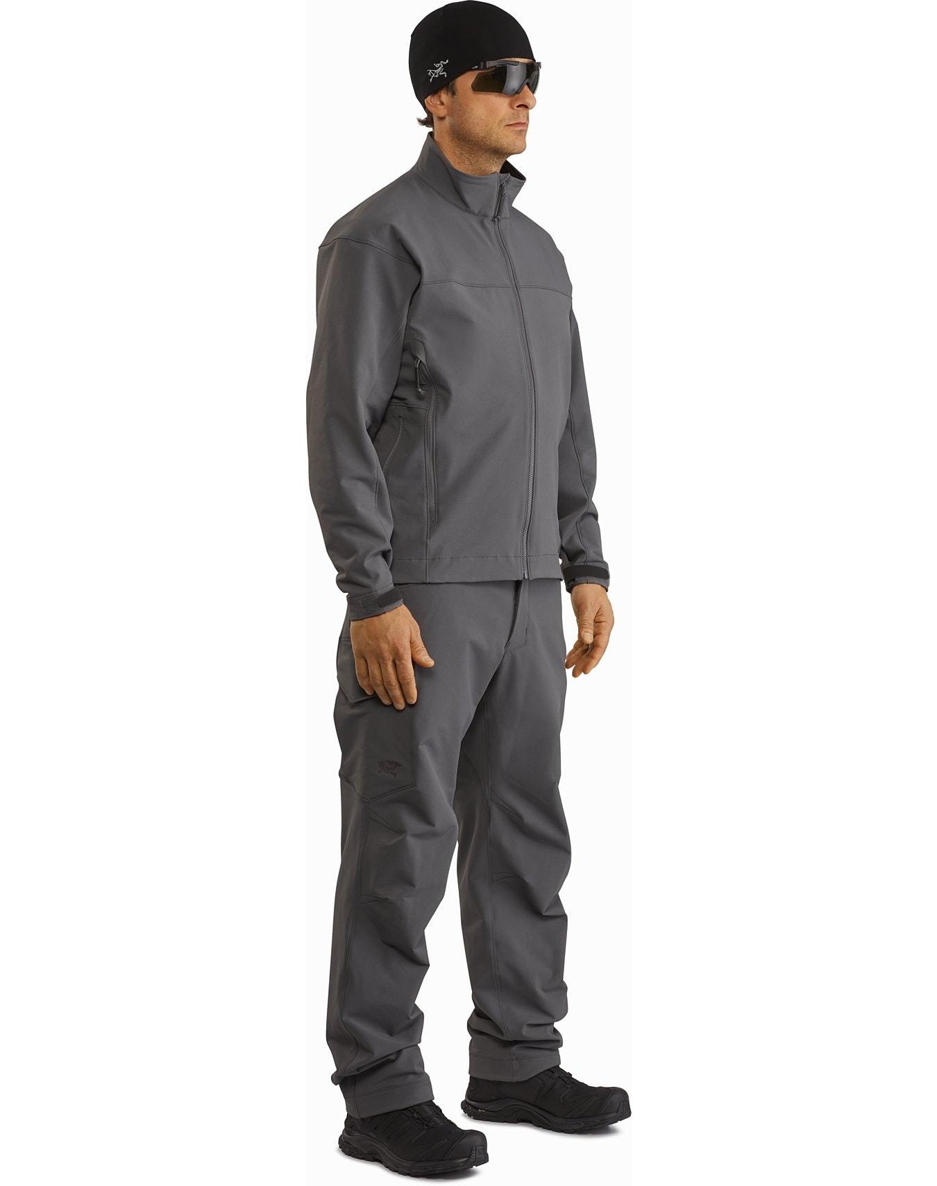 Ein Mann steht leicht gedreht, bekleidet mit schwarzer ARC'TERYX LEAF Arc'teryx Patrol Jacket AR Men's, passender Hose, schwarzen Schuhen, Sonnenbrille und einer Beanie, die an polizeidienstliche Uniform erinnert.