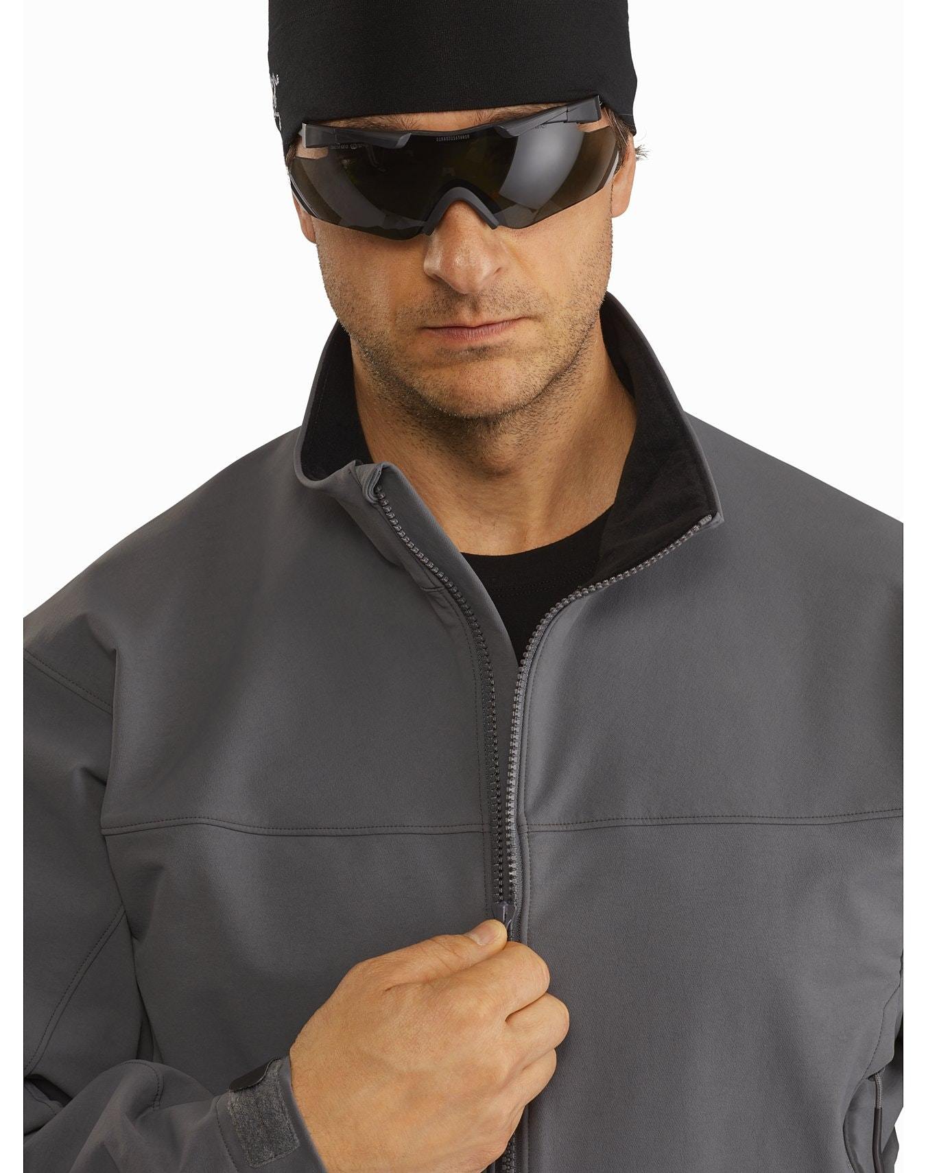 Ein Mann mit schwarzer Mütze, dunkler Sonnenbrille und einer ARC'TERYX LEAF Arc'teryx Patrol Jacket AR Men's - Wolf, einer robusten, wasserabweisenden Softshelljacke, blickt von der Brust aufwärts nach vorne, eine Hand zieht den Reißverschluss hoch.
