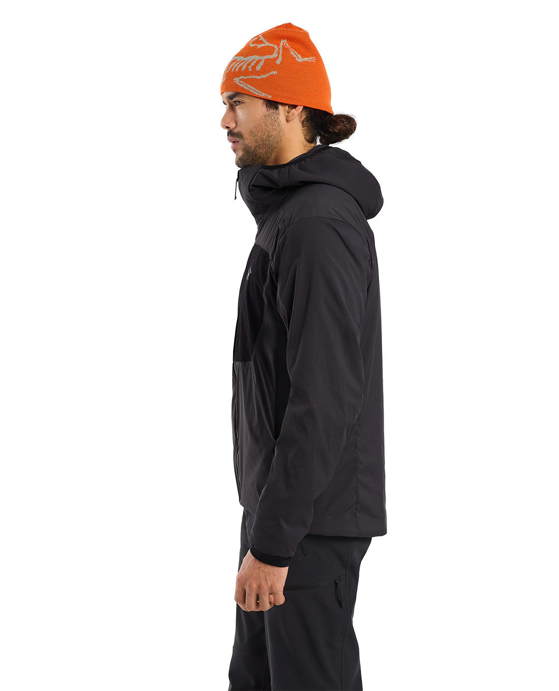 Eine Person in einem ARC'TERYX PRO Arc'teryx Practitioner AR Hoody Men's - Carbon Copy, einer schwarzen Hose und einer orangefarbenen Mütze mit weißem Muster steht im Profil vor einem schlichten weißen Hintergrund.